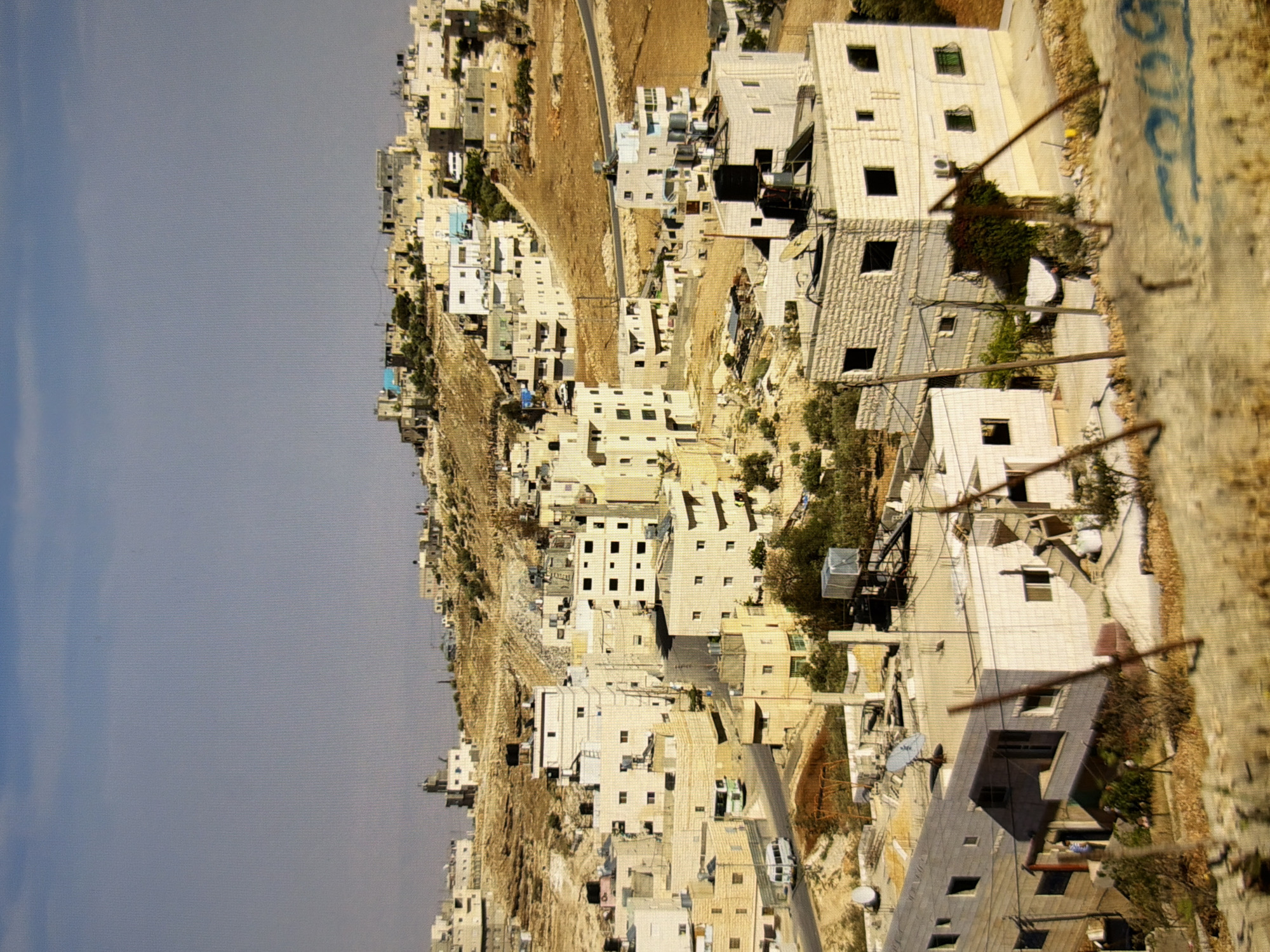 The image depicts a dense urban settlement situated on a hillside. The buildings are predominantly made of light-colored stone or concrete, with multiple stories and flat roofs. Many rooftops are equipped with water tanks, solar panels, and satellite dishes. The architecture suggests a contemporary Middle Eastern or Mediterranean style. Roads wind through the settlement, and several vehicles, including a white van and a green bus, are visible. The terrain is arid and rocky, with sparse vegetation. The sky is clear and blue. The image does not show any signs or indications that place it in Kyiv, Ukraine.