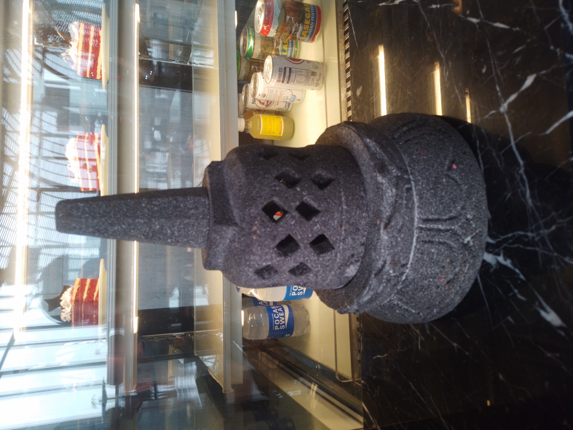 In Bumirejo, Indonesia, a dark gray, stone stupa-like object is displayed on a black marble countertop. The stupa has intricate carvings and geometric cutouts. Behind it, a glass-fronted display case reveals shelves stocked with various items. On the top shelf, slices of red velvet cake are visible. The middle shelf holds bottles of "POCARI SWEAT" and other drinks, including canned beverages. The environment appears to be a commercial establishment, likely a cafe or restaurant, with reflective surfaces and interior lighting contributing to the overall ambiance.