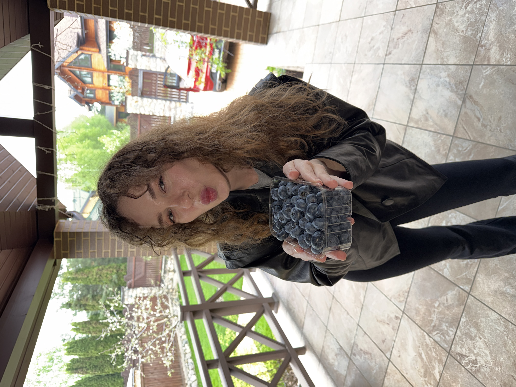 A woman with curly brown hair is holding a container of blueberries. She is wearing a dark jacket and pants. The background shows a building with brown and stone elements, greenery, and a wooden railing. The photo was taken on a tiled floor in Новоглаголево, Russia.
