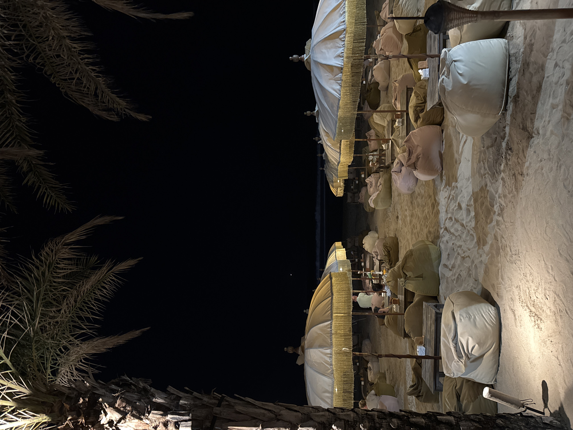An outdoor seating area is set up on a sandy beach at night, likely in Kuta, Indonesia, indicated by the presence of palm trees and the ocean. Numerous beanbag chairs and low tables are arranged under large, ornate umbrellas. Several people are seated, some at tables and others on the beanbags, appearing to be dining or relaxing. The scene is illuminated by soft lighting, casting a warm glow on the sand and the beach furniture. The ocean is visible in the background, with gentle waves breaking on the shore under a dark sky.