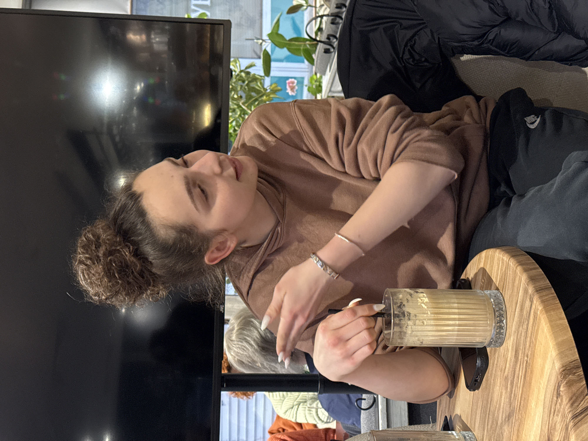 The image depicts an interior scene within a cafe or restaurant in Sofia, Bulgaria. A young female is seated at a round, light-colored wooden table with a visible grain pattern, facing partially to the right. She has dark, curly hair tied in a high bun and is wearing a brown hooded sweatshirt and dark sweatpants, which feature a white emblem on the left thigh. Her right hand is raised, fingers slightly spread and extended forward, as if gesturing, while her left hand holds a black straw in a tall, clear, ribbed glass containing a light brown, frothy beverage. She wears two silver bracelets on her right wrist and has long, white, almond-shaped fingernails. A black smartphone in a black case lies flat on the table to her left, alongside a partial view of another clear glass.
In the immediate background, a large, dark flat-screen television monitor bearing the "JVC" brand name is mounted. Reflections of ambient light and distant objects are visible on its surface. To the left of the main subject, two other patrons are partially visible: one with gray hair wearing a light green top, and another with reddish-orange hair wearing an orange garment. To the right, green potted plants are arranged near a large window. Through the window, a light blue exterior wall and a partial reflection of Cyrillic script, appearing as "УР," are visible. A black padded jacket is draped over a chair or bench to the main subject's right, next to a beige upholstered seat. The precise establishment within Sofia, Bulgaria, cannot be ascertained from the visual evidence.