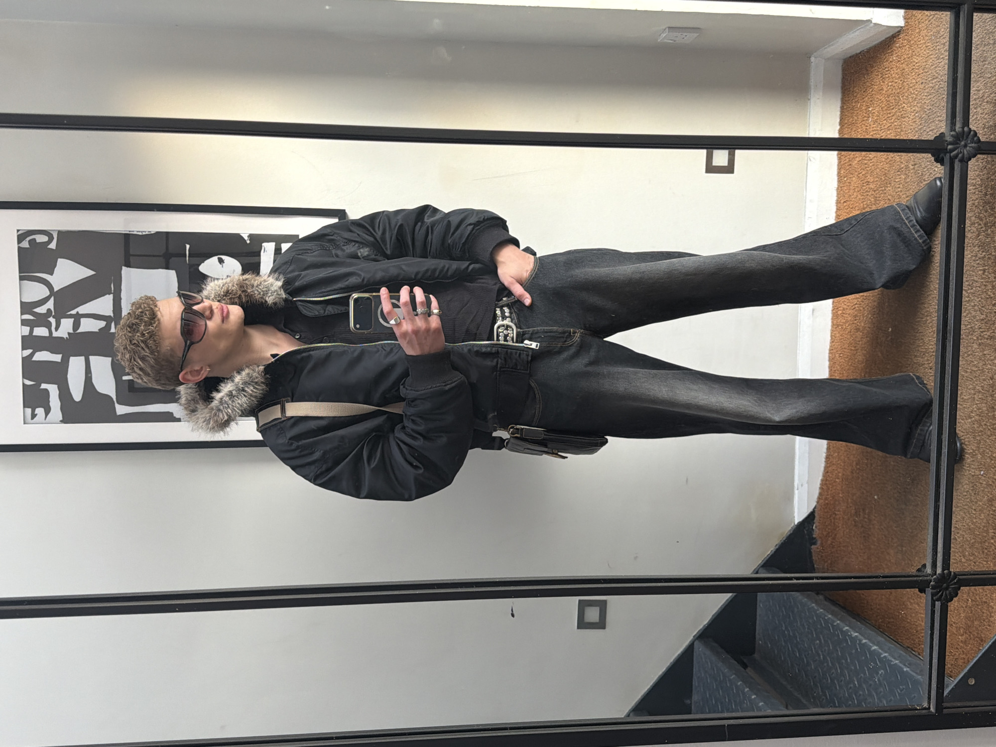 A young male with fair skin, short curly light brown hair, and dark sunglasses is posing for a photo in a mirror. He is wearing a black bomber jacket with a fur-lined collar, a black t-shirt, dark jeans, and black shoes. He has several rings on his fingers. In his left hand, he is holding a smartphone. He also wears a dark, ornate belt. A small tan shoulder bag is slung across his torso.
The mirror has a black metal frame. The background includes a white wall, a framed black and white print, carpet, a partial view of metal stairs, and light switches.
Based on the style of the building and the visible staircase, the location is likely a residential or commercial interior in London, United Kingdom.