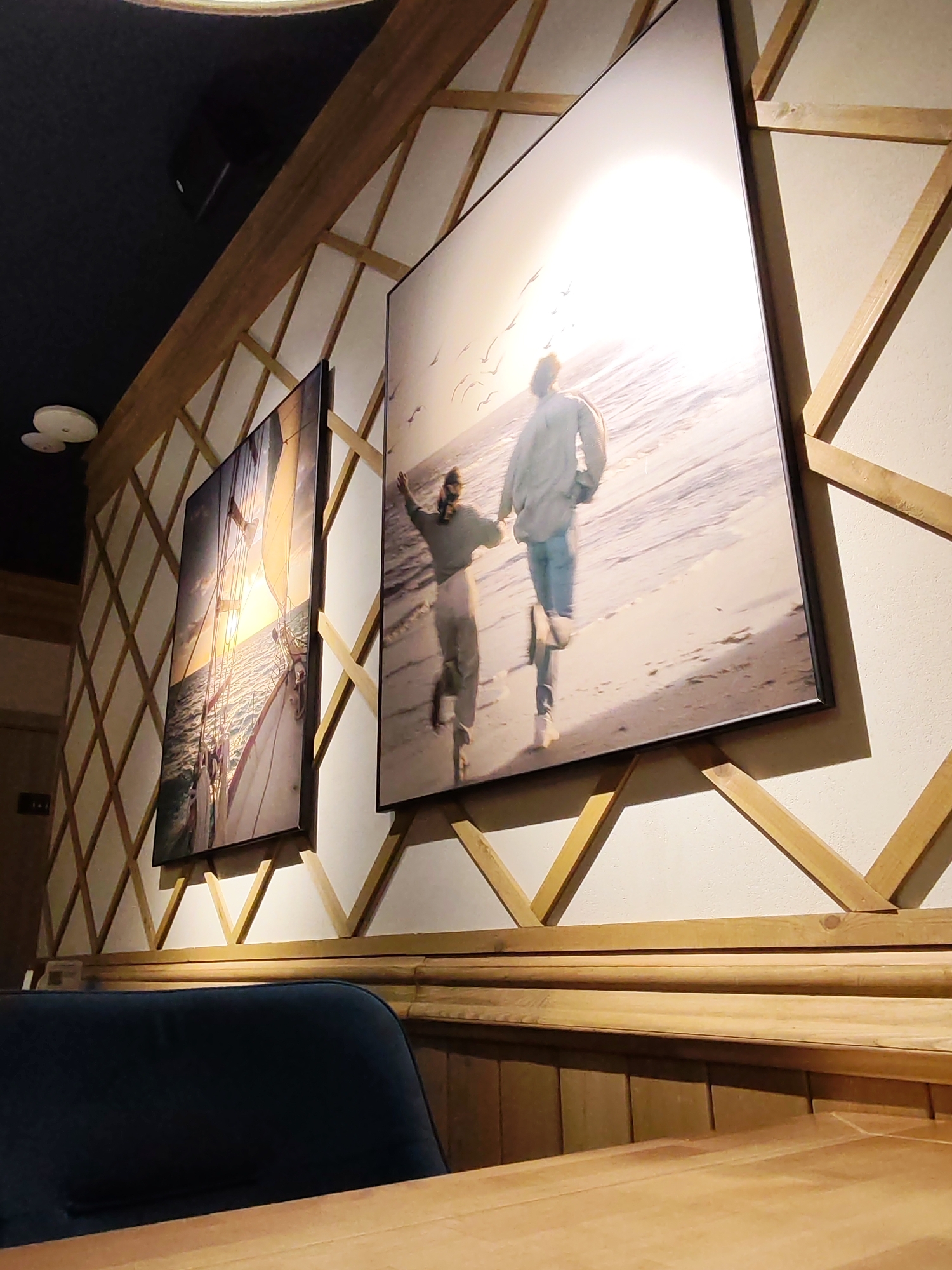 This interior shot, taken in Izhevsk, Russia, captures a section of a room, likely a cafe or restaurant, characterized by warm wooden tones and inviting imagery. The perspective is low, looking up towards a decorative wall.
The main subject is a wall featuring a striking wooden lattice pattern, where light-colored wooden strips form diagonal diamond shapes against a light-colored background. Below this decorative section, there's wooden paneling and a horizontal wooden dado rail, matching the table in the foreground. A dark blue upholstered bench or seating is partially visible on the left side, behind the wooden table. The ceiling on the upper left is dark, and a round, white light fixture is mounted on it.
Two large, framed pictures are prominently displayed on the lattice wall. The picture on the left shows a sailboat on the water, bathed in the golden light of either a sunrise or sunset. The sun is low, casting a warm glow across the scene. The picture on the right depicts two people, appearing to be a couple, running hand-in-hand along a sandy beach. They are back-lit by a very bright sun, again suggesting sunrise or sunset, with several birds visible in the bright sky above them.
The activity within the pictures portrays moments of freedom, romance, and natural beauty—sailing and running on a beach. No specific activity or event is taking place in the immediate room itself, which appears to be set for patrons. The notable details include the consistent warm lighting and natural themes in the artwork, evoking a serene and comfortable atmosphere. No visible text is present in the image.