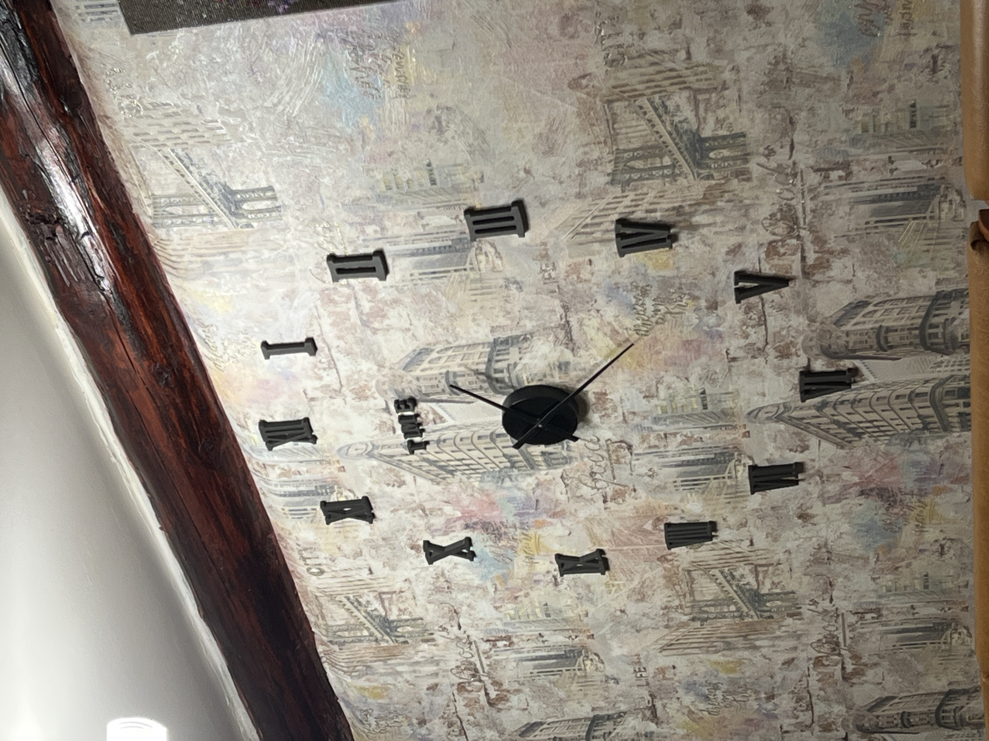 A photograph taken indoors shows a wall clock with Roman numeral markings. The clock hands indicate a time around 10:20. The wall has a textured, patterned surface with the look of faded newsprint or architectural drawings. A dark wooden beam runs along the left side of the image, suggesting the presence of a structural element or decorative feature in the room. There is no indication of the image being taken in Сосново, Russia.