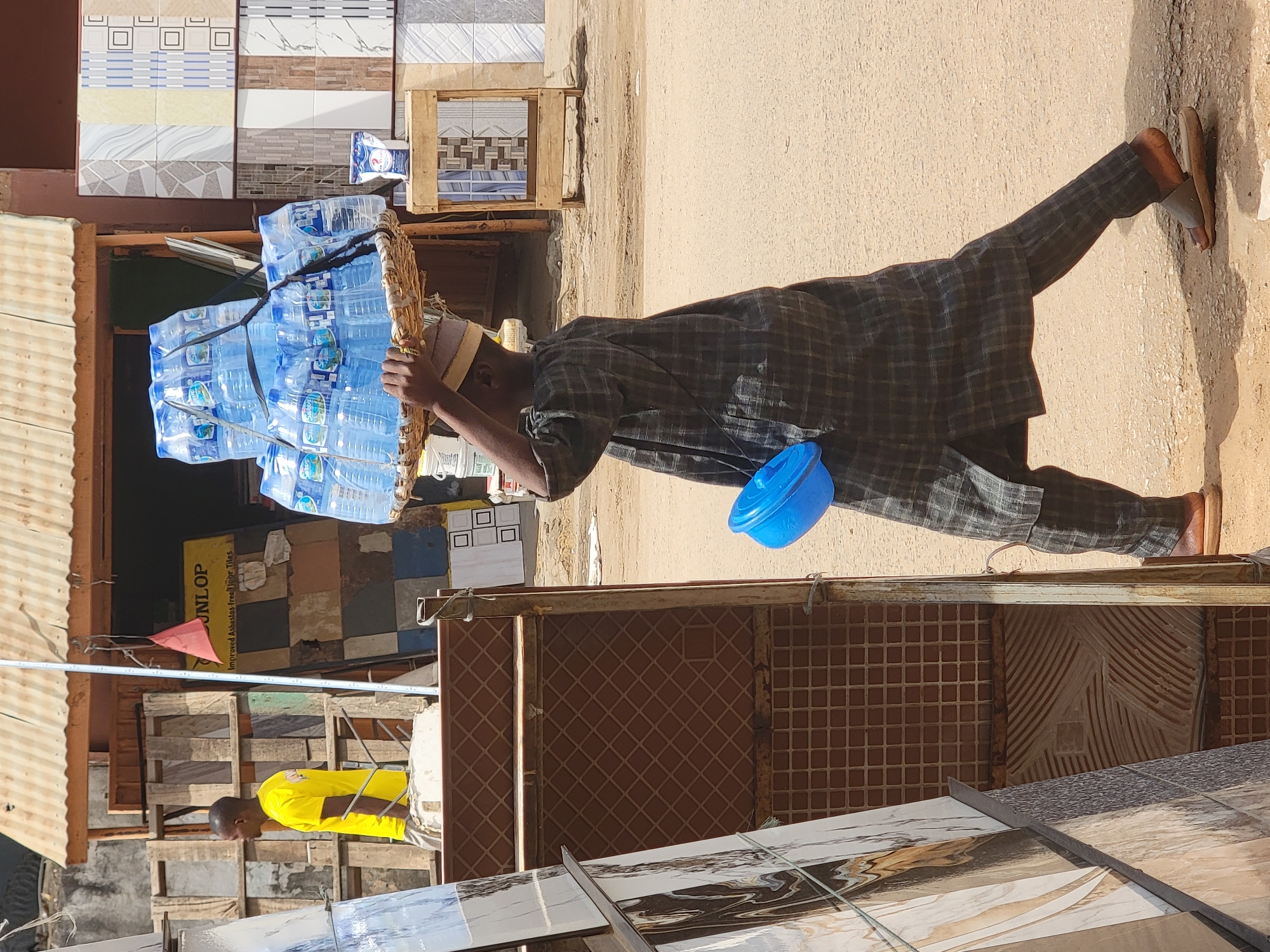 A man wearing a dark checkered shirt and pants, as well as a white cap, is carrying a basket piled with water bottles on his head. He is walking on a dirt or paved surface beside a building displaying tiles for sale. A blue bowl hangs from his side. A second man in a yellow shirt is visible in the background. The location appears to be a commercial area in Maiburiji, Nigeria.
