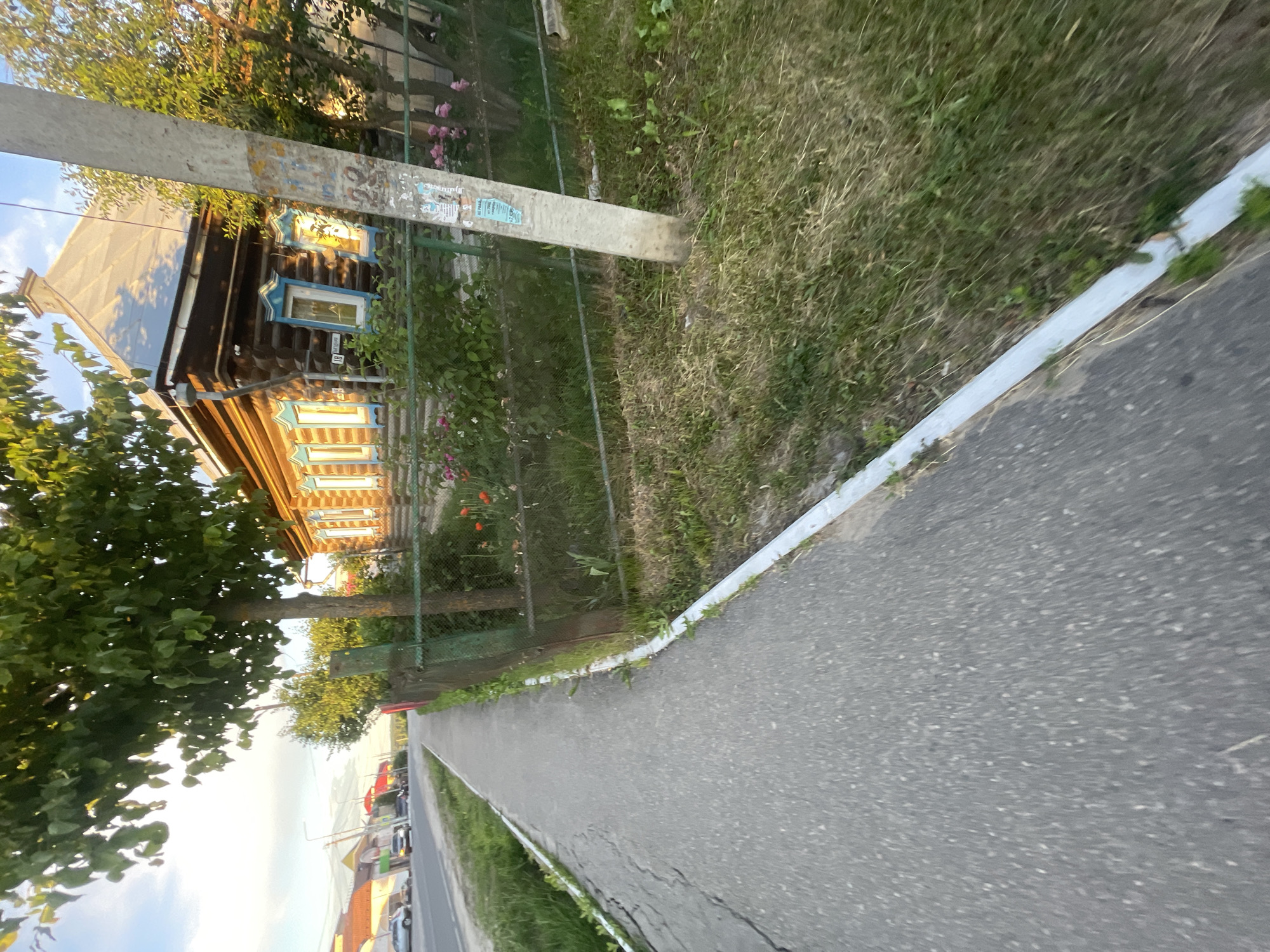The image depicts a sidewalk in the city of Krasnye Baki, Russia, on the left side of the frame. The sidewalk is made of asphalt and has a concrete curb. To the right of the sidewalk is a grassy area with a metal fence. Beyond the fence is a traditional wooden house with blue window frames and a peaked roof. The house appears to be well-maintained. There are trees and bushes surrounding the house, and some flowers are visible in the garden. A utility pole stands near the fence, with various notices attached to it. In the background, a road with cars and buildings can be seen, suggesting that this is a residential area within the city. The sky is visible at the top of the frame, with clouds and sunlight indicating it is daytime, likely late afternoon or early evening due to the warm lighting.