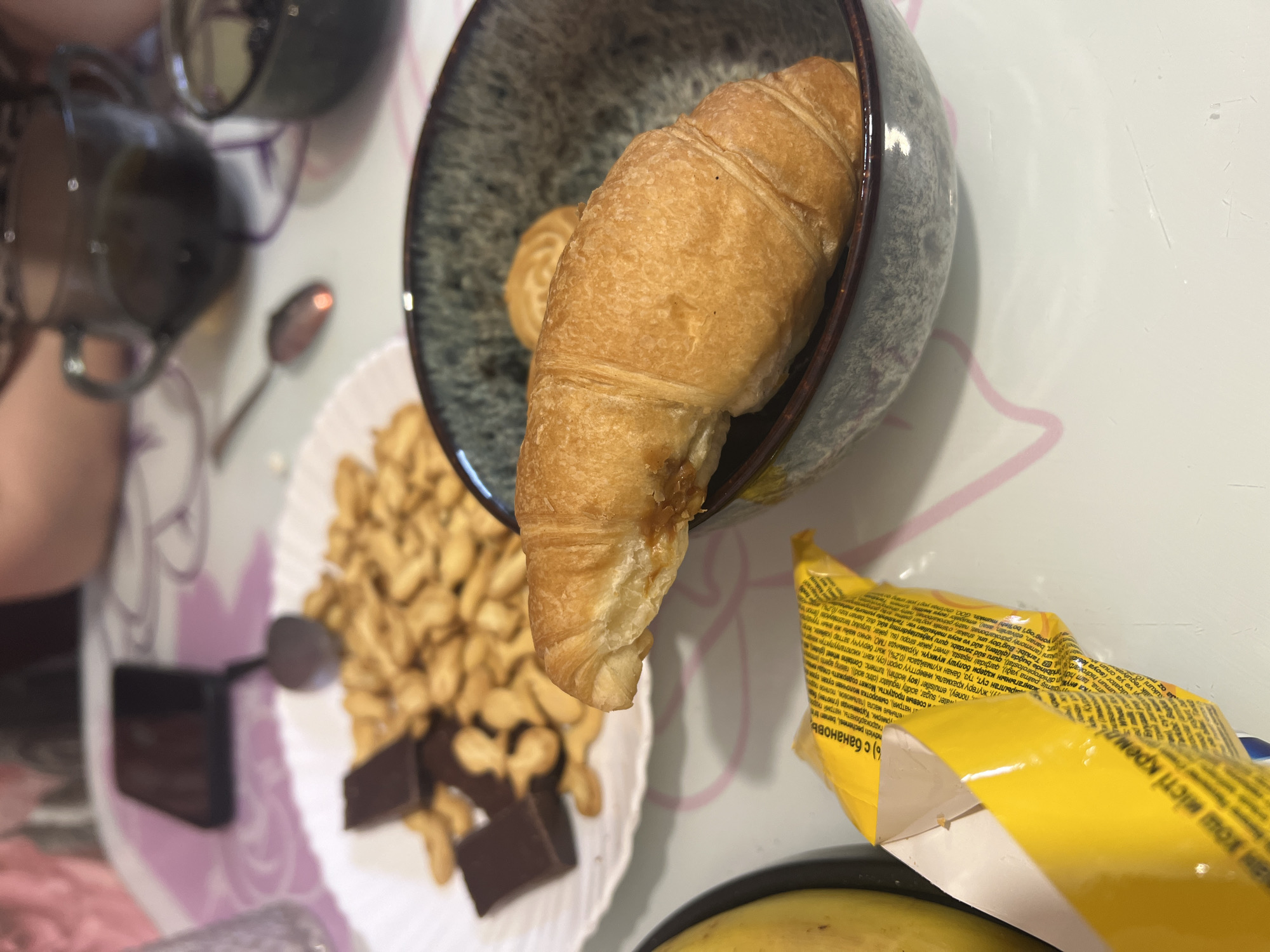 A still life arrangement presents an overhead view of breakfast or snack items on a table. A croissant rests in a speckled gray-blue bowl. A white plate holds a pile of cashew nuts, two pieces of dark chocolate, and a small black object (possibly a USB drive or other small device). There is an open yellow wrapper from a snack that is printed in Russian, resting near the plate. A banana is visible beneath the wrapper. Reflections of two dark cups are seen in the top left, with two spoons also being visible.