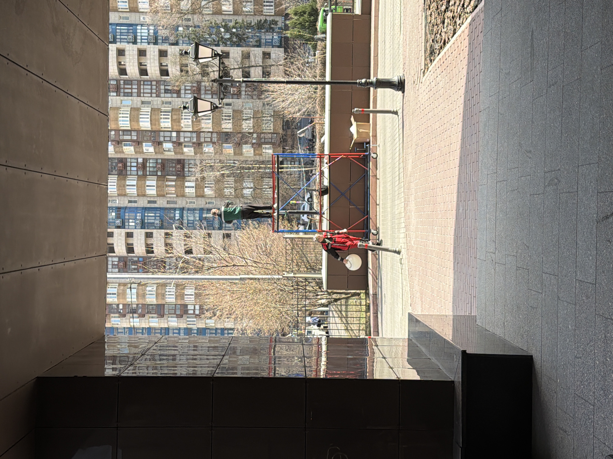 The image shows an outdoor urban scene, likely in Moscow, Russia, based on architectural cues and style. The photograph is taken from a shadowed alcove formed by a building or structure. Outside the alcove, the scene is brightly lit. A multi-story building with numerous windows is in the background, with its architecture hinting at a post-Soviet, modern style.
In the middle ground, there is a street with a lamppost and scaffolding. One man is on the scaffolding wearing a green jacket and another man is standing on the ground next to the lamppost, wearing a red uniform. There are construction materials near the scaffolding.
The ground is paved with tiles of varying colors, with a clear distinction between the paving inside the alcove and the external street. The image orientation is rotated 90 degrees counter-clockwise.