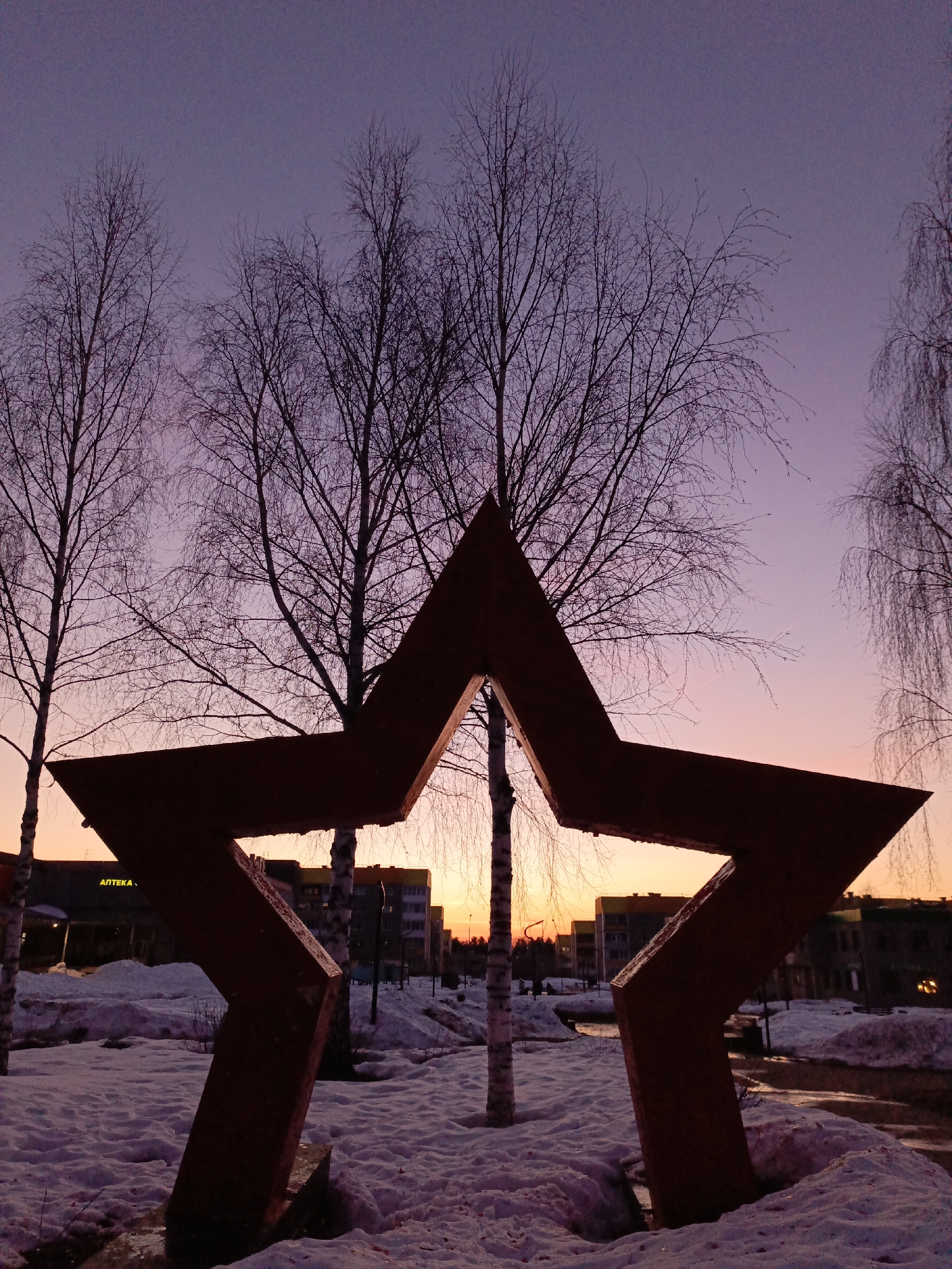 The image captures a public park scene in Markovskiy, Russia during twilight. The foreground features a large, rusty red star sculpture partially buried in snow. Several bare birch trees are positioned behind the star, their branches stark against the fading sky, which transitions from purple to orange. The middle ground shows snow-covered ground with patches of water, reflecting the dim light. In the background, a row of apartment buildings is visible, with a pharmacy sign displayed on one. There are no people visible in the image. The overall atmosphere is cold, quiet, and indicative of a late winter or early spring setting.