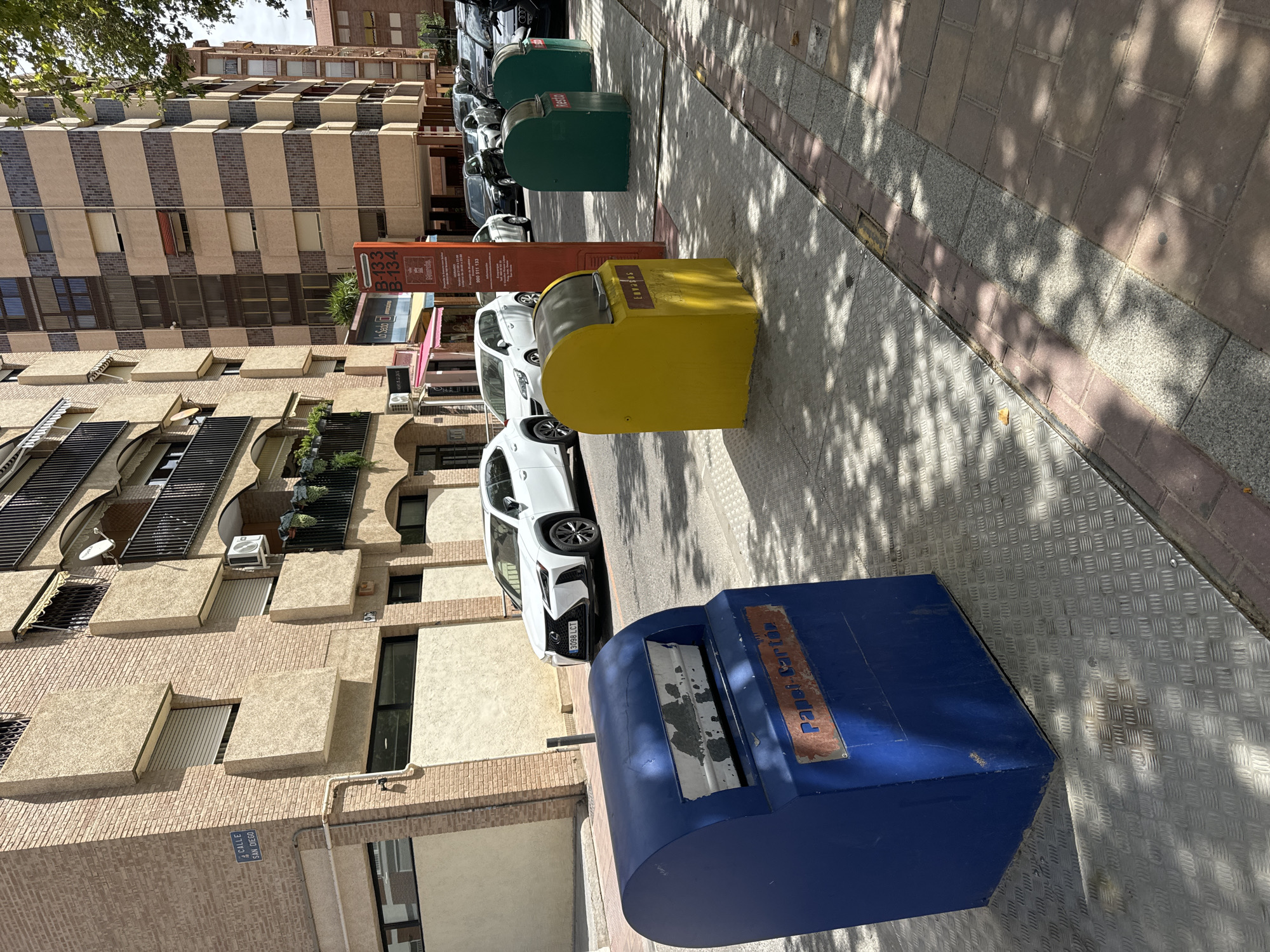 The image depicts a street scene in Murcia, Spain, with a focus on a row of recycling bins. In the foreground, a large blue bin labeled "Papel-Cartón" (Paper-Cardboard) is prominent. To its right, a yellow bin labeled "ENVASES" (Containers) and two green bins labeled "RESTO" (Waste) are visible. The bins are situated on a metallic, textured surface adjacent to a sidewalk paved with bricks and granite. In the background, white and dark-colored cars are parked along the street. Behind the cars, there are residential buildings with balconies and awnings. A street sign visible in the upper left corner reads "CALLE SAN DIEGO" (San Diego Street). The scene is bathed in sunlight, casting shadows from trees onto the ground. There are no people visible in the image.