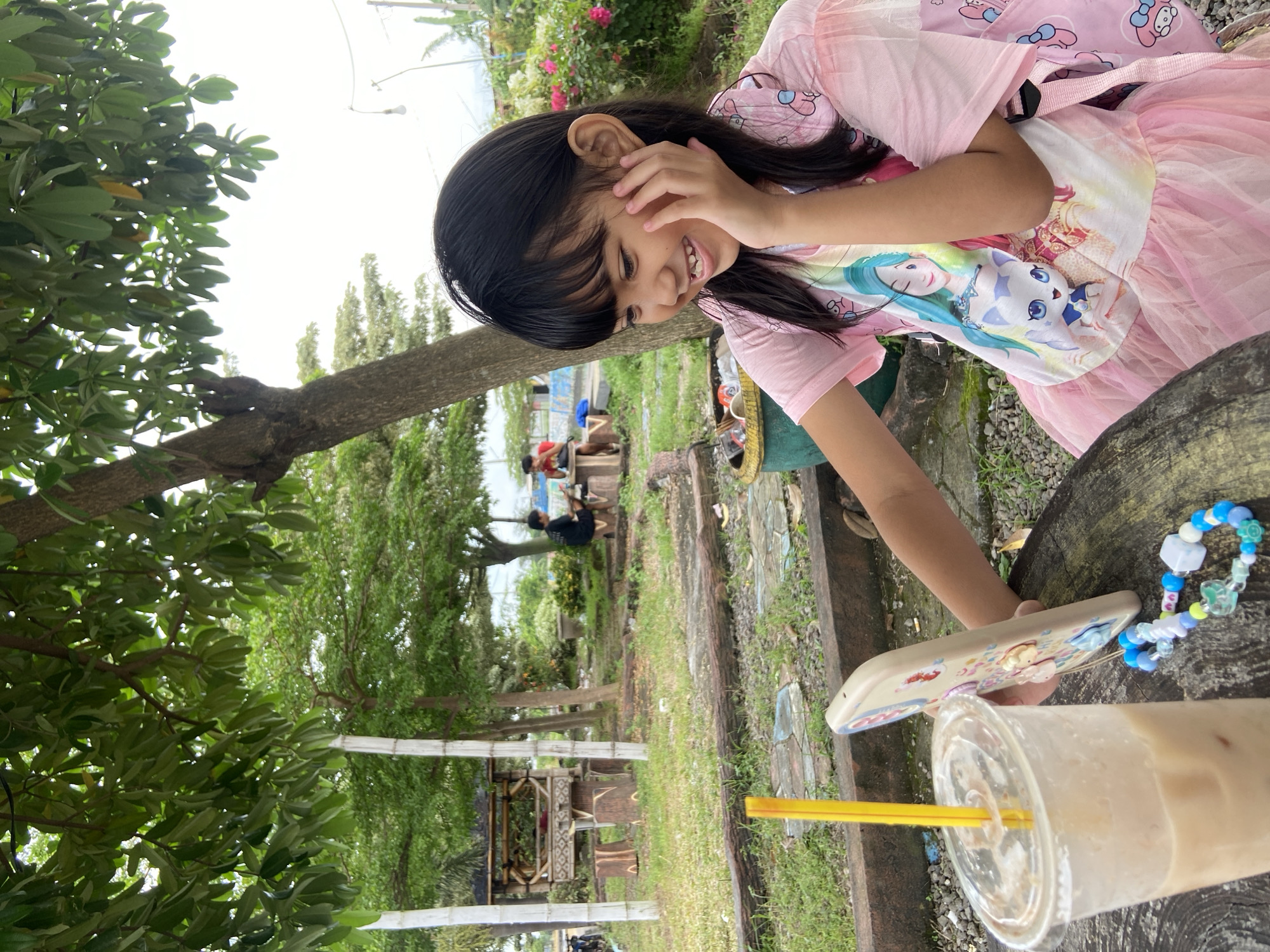A young girl is depicted seated outdoors, likely in a park or garden setting in Kalitengah, Indonesia. She is wearing a pink dress with a print of animated characters and a matching backpack. Her right hand is touching her face while she looks down and smiles. In front of her is a clear plastic cup with a straw, possibly containing a milky beverage. Beside it is a mobile phone in a decorative case and a beaded bracelet. The foreground includes a rough wooden surface, possibly part of a tree stump used as a table. The background features trees, other greenery, and in the distance, people are sitting on benches.