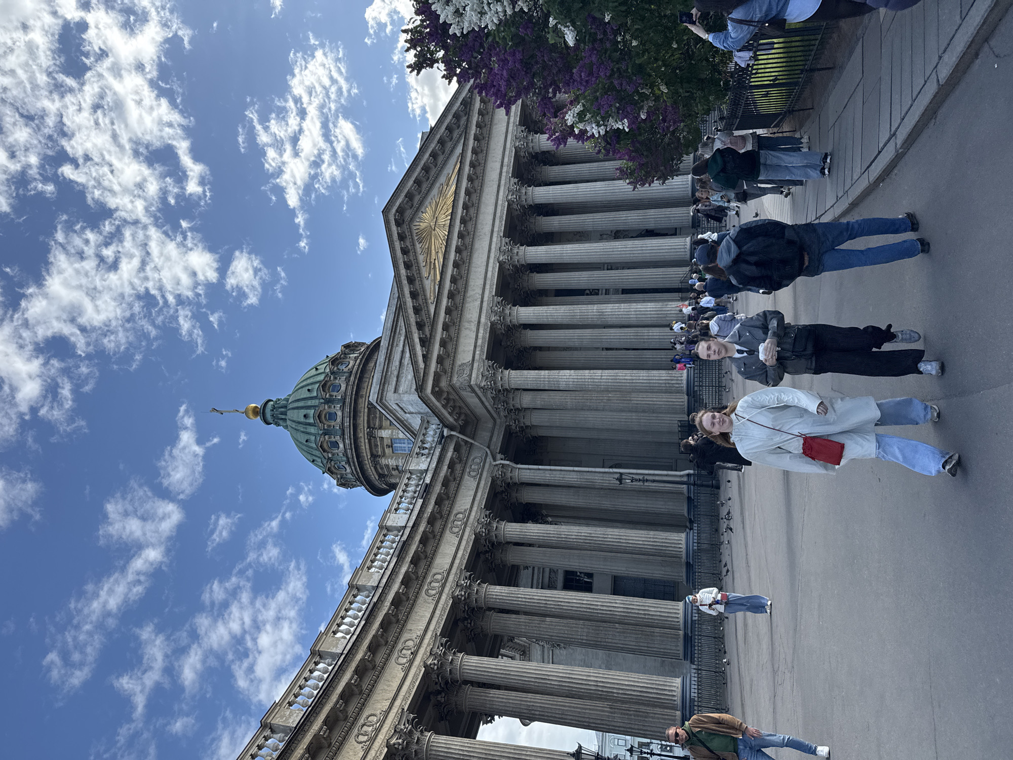 The photograph depicts the Kazan Cathedral in Saint Petersburg, Russia, framed against a sky with scattered clouds. The architectural details of the cathedral, including its dome, colonnades, and facade, are visible. Several people are walking on the pavement in front of the building. Lilac branches are visible in the upper part of the frame.