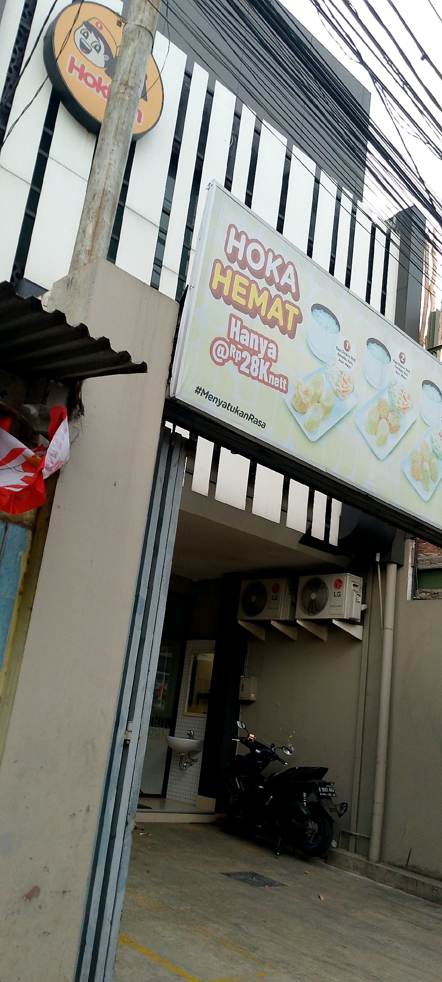 This is a medium-shot, angled exterior shot of a commercial building in Bandung City, Indonesia. The building has a modern facade with white vertical slats. A circular yellow sign with a cartoon face and the word "Hoka" in red is prominently displayed on the left side of the building. Below it, a large rectangular signboard advertises "HOKA HEMAT HANYA @Rp28Knett #MenyatukanRasa" with images of food.
The foreground is dominated by a concrete pillar and a corrugated metal awning. A motorcycle, dark in color, is parked on the right side of the frame, facing towards the building. In the background, two LG air conditioning units are mounted on the wall. Through an open doorway, a sink and a tiled wall are visible. The ground is concrete, with yellow lines painted on it. Overhead, a tangled mass of electrical wires stretches across the frame. There are no people visible in the scene.