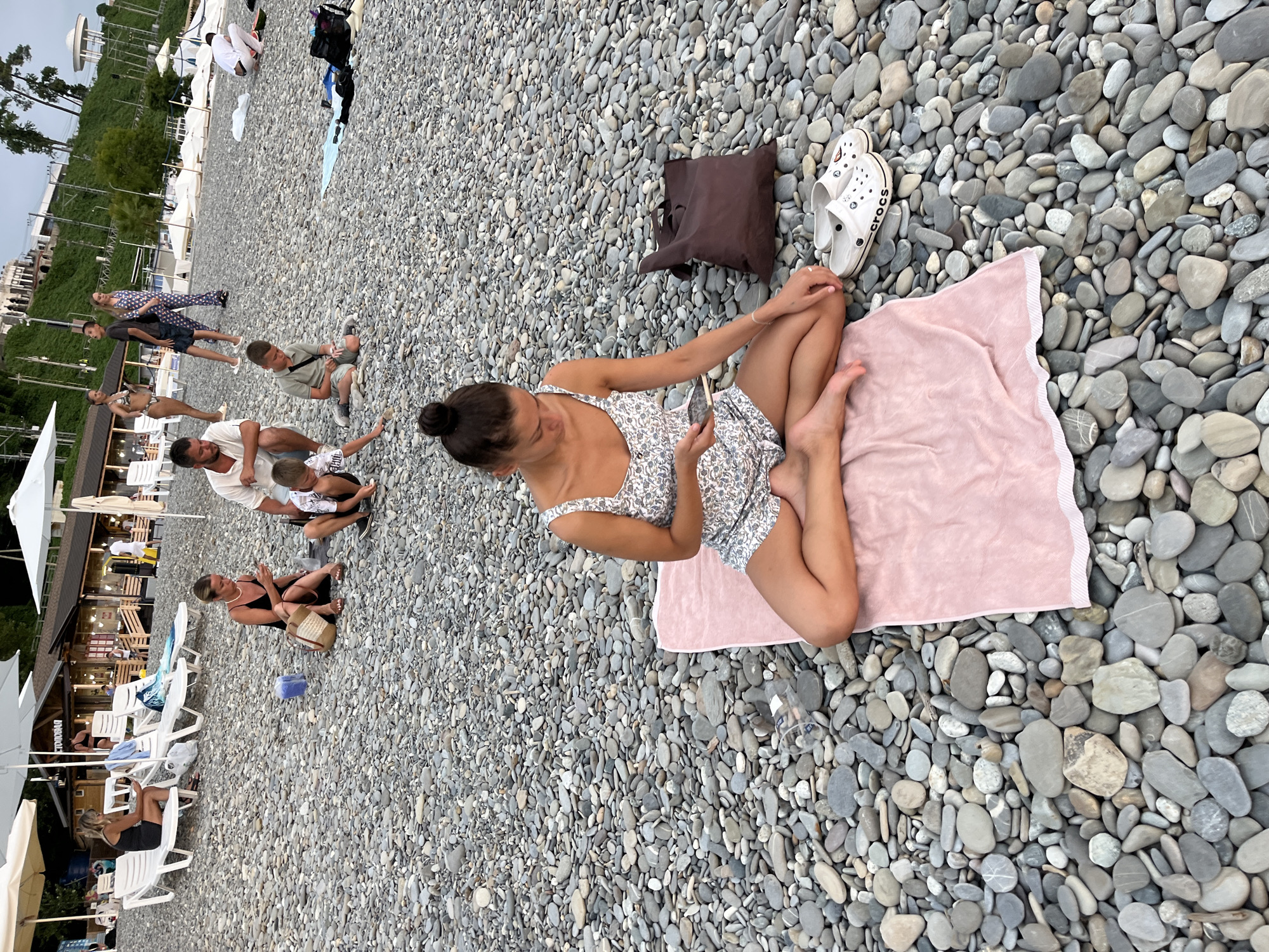 A scene unfolds on a pebbled beach in Sochi, Russia, featuring several groups of people relaxing and interacting. In the foreground, a young woman with her hair in a bun is seated cross-legged on a pink towel, looking at her phone. Beside her are a brown tote bag and a pair of white Crocs.
Further back, a family is gathered on the beach. A man in a white shirt is crouched down, facing away from the camera. A boy in a grey t-shirt is sitting nearby, also looking at his phone. Another boy in a patterned shirt is closer to the man.
In the middle ground, two women are seated on lounge chairs, each looking at their phones. One woman wears a black dress, and the other a patterned bikini. Behind them, a building with outdoor seating and umbrellas suggests a cafe or restaurant.
In the background, more beach umbrellas and lounge chairs are visible, along with people strolling along the shore. The terrain is covered in smooth, grey pebbles, with the sea likely out of frame. The overall atmosphere is one of a relaxed day at the beach.