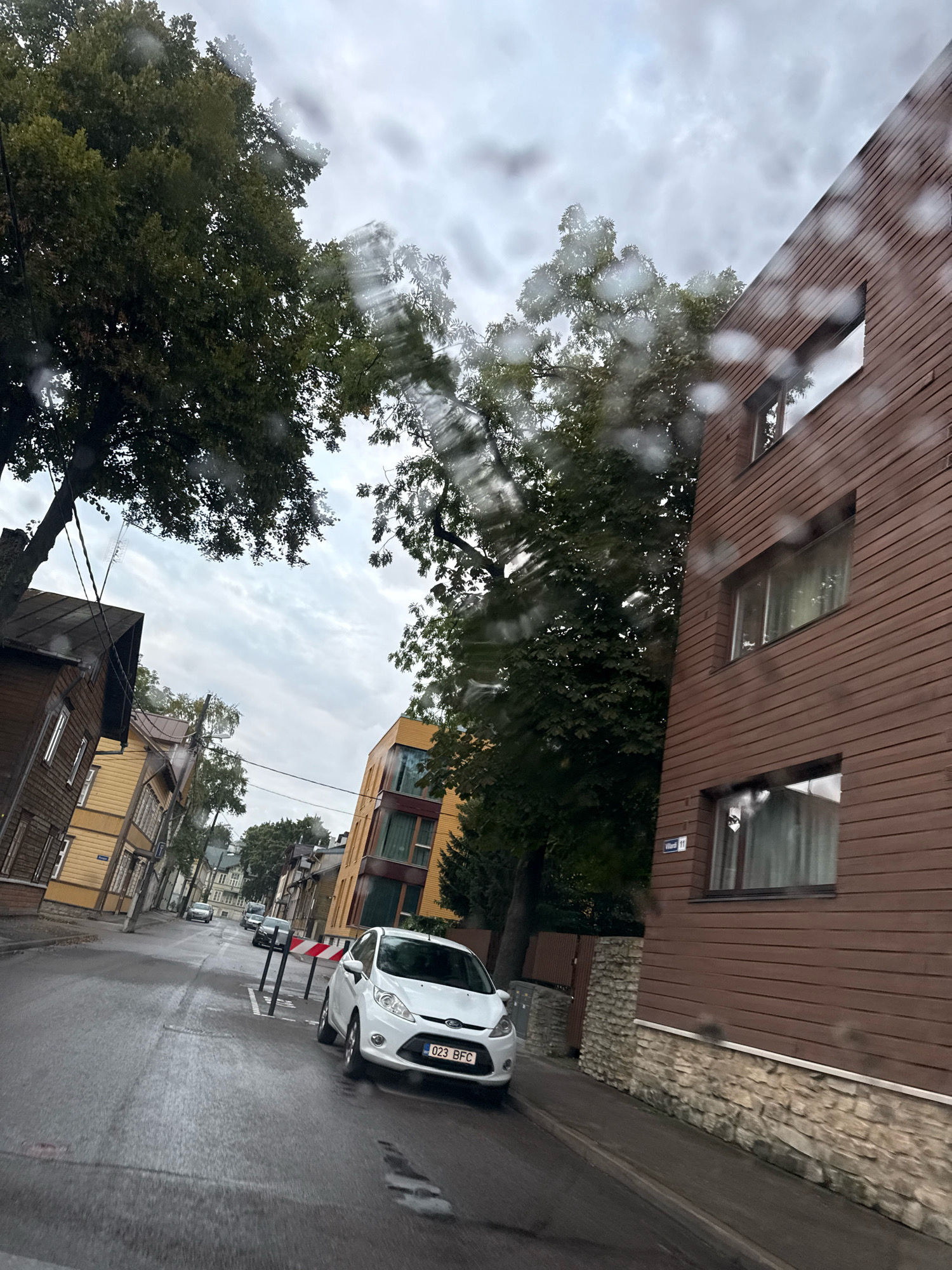 The image depicts a street in Tallinn, Estonia, on a cloudy day. The street is wet, suggesting it might be raining or has recently rained, as evidenced by raindrops on the camera lens. The perspective is from inside a vehicle, looking forward and slightly to the right.
On the right side of the street, a modern building with brown wooden siding and multiple windows dominates the foreground. The building has a visible address sign that reads "Välja 11". Adjacent to it is a stone foundation.
On the left side of the street, there are older, wooden houses painted in yellow and brown. These houses appear to be typical of older European architecture.
A white Ford Fiesta car with the license plate "023 BFC" is parked on the right side of the street. Further down the street, several other vehicles are visible, including a dark-colored car and a few other cars parked along the side. A temporary traffic barrier with red and white stripes is placed in the middle of the road.
Trees with green foliage line the street, contributing to the urban landscape. The sky is overcast with a mix of gray clouds and patches of lighter sky.