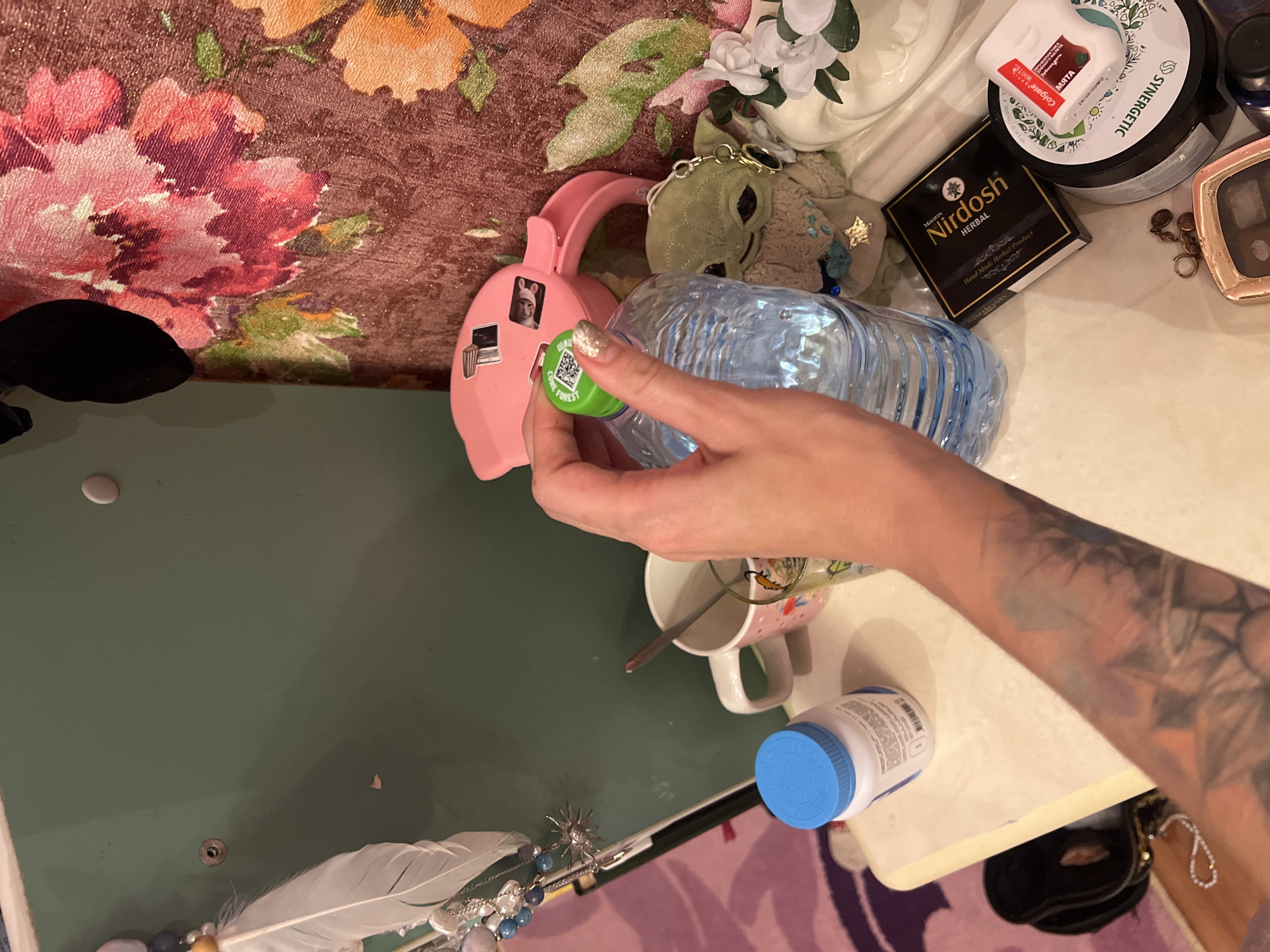 In Krasnoe-na-Volge, Russia, a person's hand with glitter nail polish is holding a plastic water bottle with a green cap. The bottle has a QR code on the cap. Next to the bottle, there is a small plush toy resembling Grogu from Star Wars, a pink electronic device, a mug with a spoon, a white bottle with a blue cap, a black handbag, and various cosmetic products including a box labeled "Nirdosh Herbal" and a container with "SYNERGETIC" written on it. The background features a textured wallpaper with floral patterns in shades of pink, orange, and brown, and a green wall. The scene appears to be on a dresser or table, with a patterned rug visible in the lower left corner.