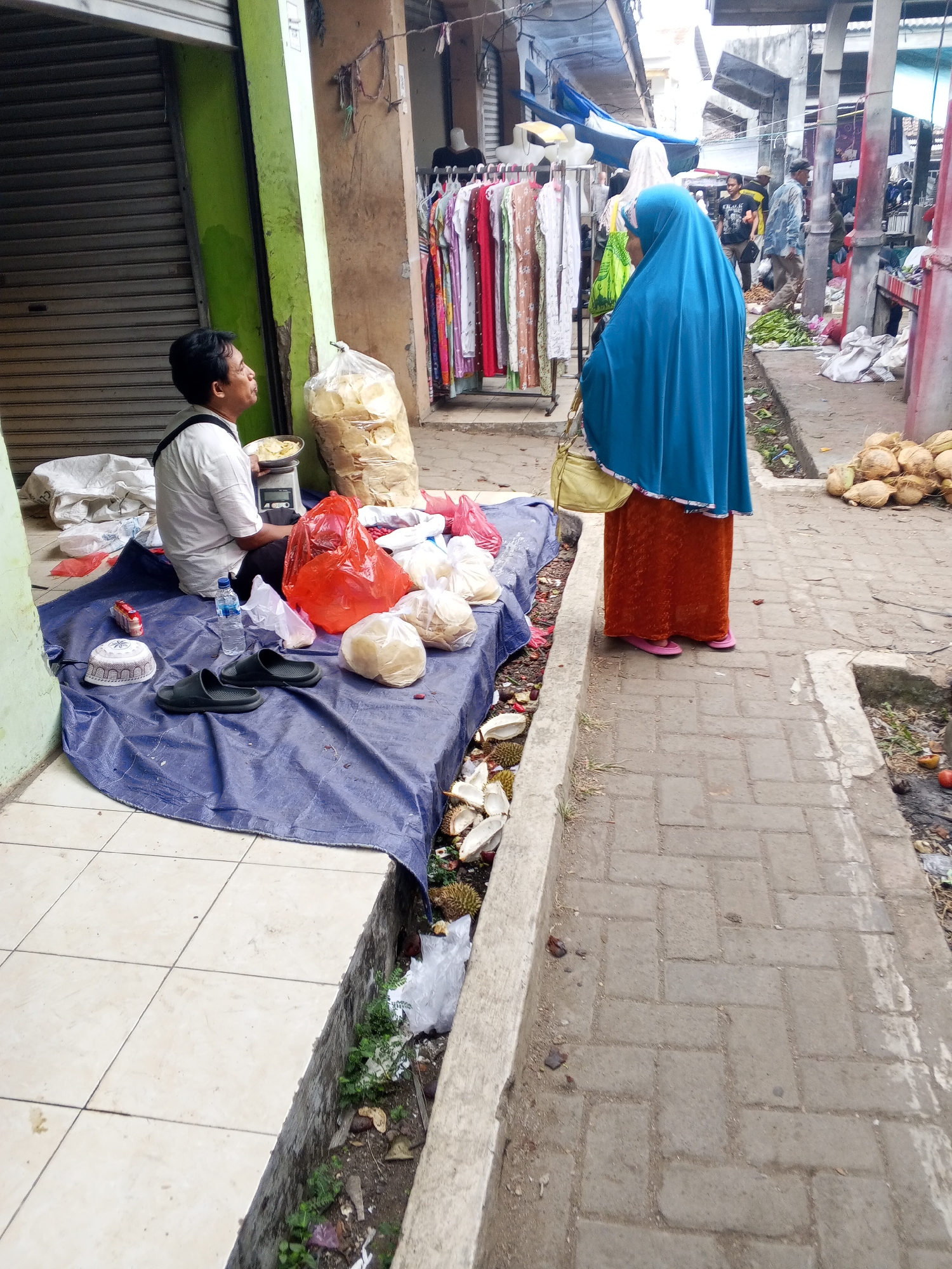The image depicts a street market scene in Serang, Indonesia. In the foreground, a man in a white shirt and black pants sits on a blue tarp, his back to the viewer. He is holding a plate and appears to be weighing something on a scale. Around him are various items, including bags of food, a blue water bottle, a pair of black sandals, and a white and patterned prayer cap. A woman in a bright blue hijab and an orange skirt stands facing away from the camera, looking towards the stalls. In the background, more market stalls are visible, with racks of clothing and other goods. The ground is a mix of tile and brick pavement, with some debris and vegetation visible along the edges. Coconuts are piled on the right side of the image, suggesting a possible vendor of fruit. The overall impression is one of a busy, open-air marketplace.