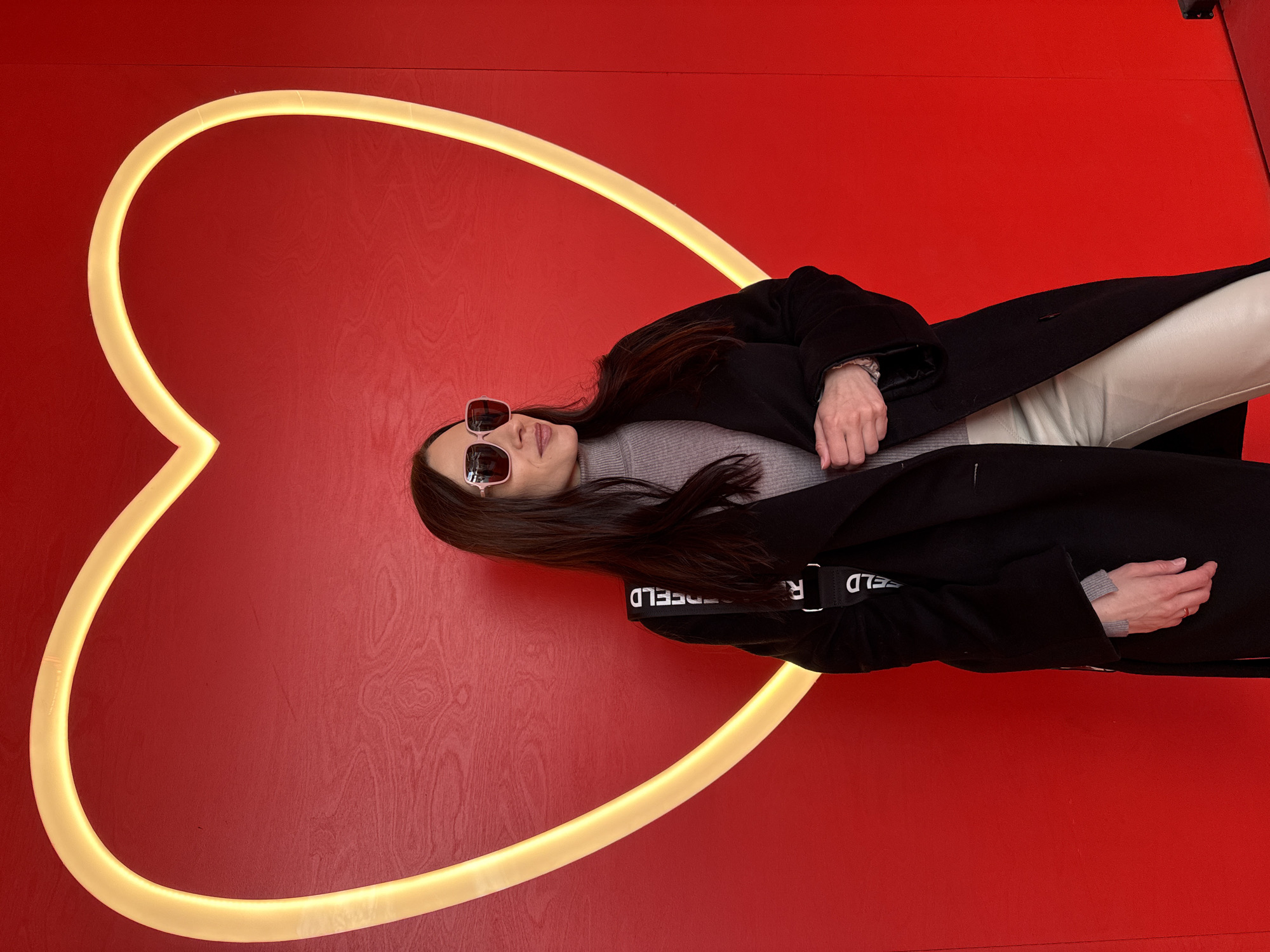 A person is standing in front of a large heart-shaped light on a red wall. The person is wearing sunglasses, a gray shirt, a black jacket, and light-colored pants. The jacket has the word "KARL" on the shoulder strap. The location is Saint Petersburg, Russia.