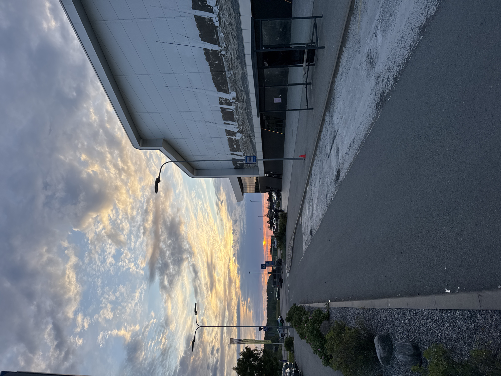 The image captures a twilight scene in Tallinn, Estonia, likely near a commercial area. The sky is a dramatic canvas of clouds, illuminated by the setting sun, casting warm hues of orange and yellow against streaks of blue and grey. In the foreground, an asphalt road curves gently to the left, bordered by a gravel and planted median strip with large rocks. To the right, a modern building with a white, textured facade dominates the scene. A large mural depicting sailboats on water adorns the building's exterior. Adjacent to the building is a bus stop shelter with a metal frame and glass panels. A blue bus stop sign is visible near a lamppost. Several cars are parked along the roadside. The overall impression is one of an urban periphery at dusk, with architectural elements contrasted against the natural beauty of the sky.