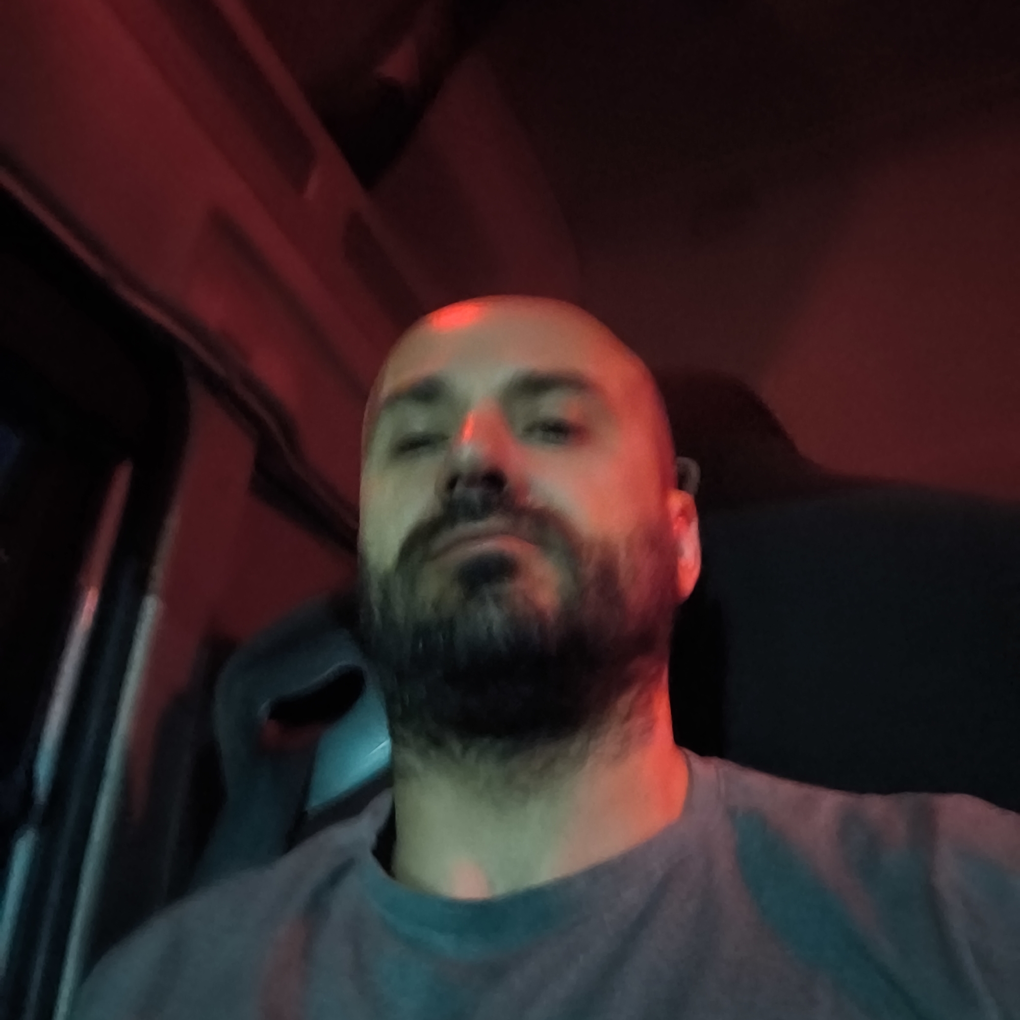 A close-up, blurry photo depicts a bald man with a beard wearing a gray t-shirt. The lighting is predominantly red, with some teal. The setting appears to be inside a vehicle, possibly a truck, with a dark interior and part of a driver's seat visible. The photo's angle is from below, looking up at the man. There is no clear indication of a location within Alcàsser, Spain, or any specific activity or interaction taking place.