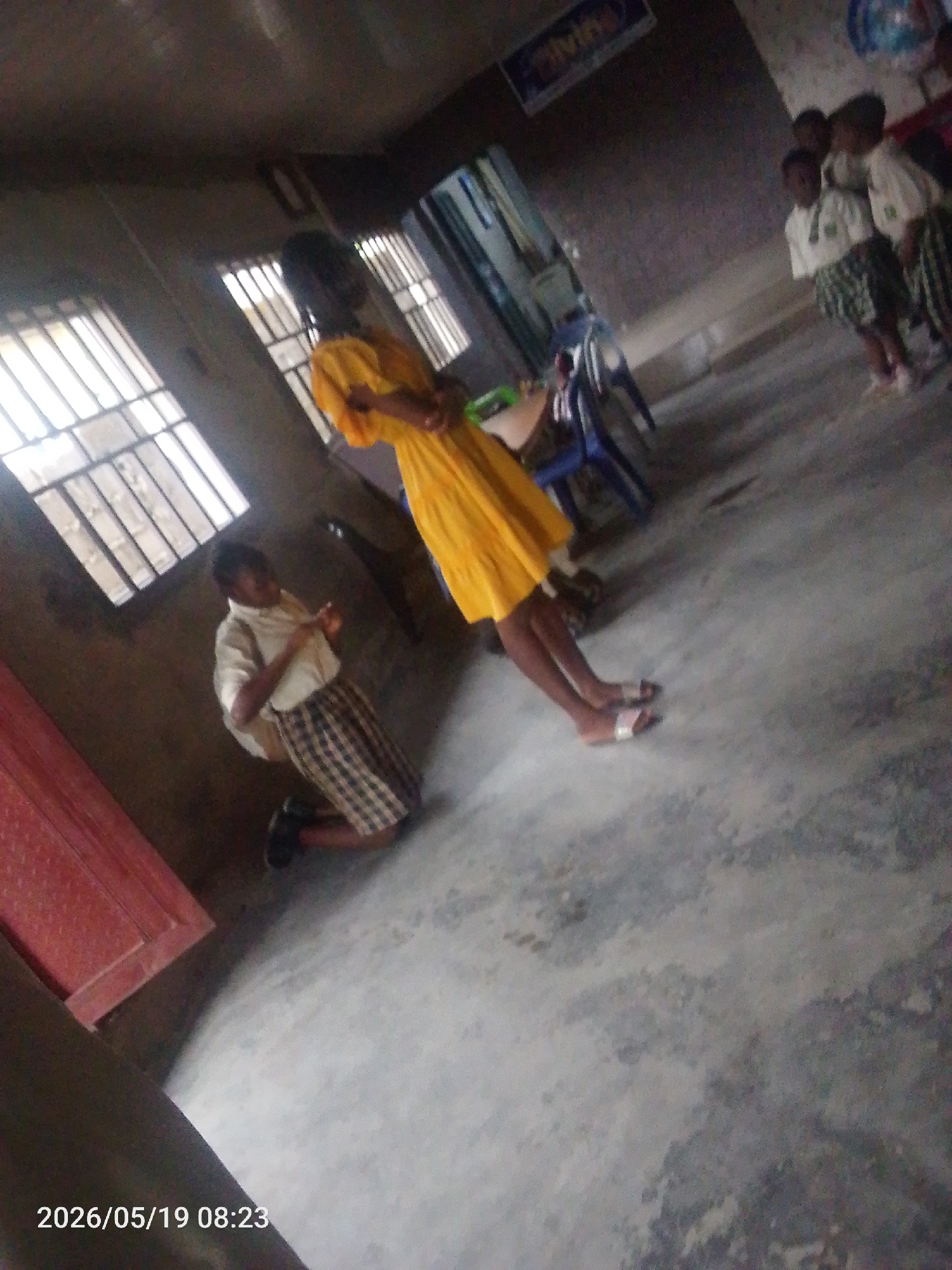 The image depicts a scene inside what appears to be a school classroom in Lokoja, Nigeria. A girl in a yellow dress is standing in the center of the room. Near the window, a girl in a school uniform kneels on the floor. A group of other students stand in the background, also in school uniforms. The classroom has a weathered appearance.