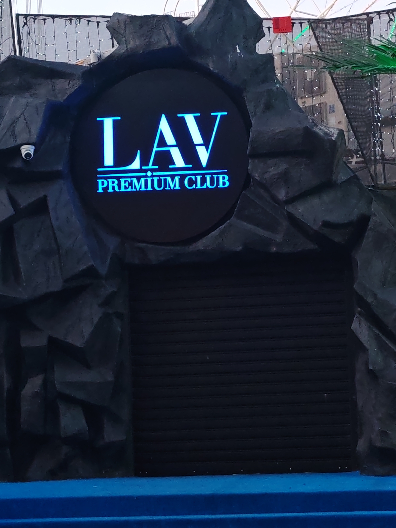 The image shows the entrance of the LAV Premium Club, presumably located in Nesebar, Bulgaria. The entrance is framed by a dark, rocky structure with a circular sign displaying the club's name in blue lettering. A security camera is visible on the left side of the structure. The entrance is closed, with a dark, horizontally striped roller shutter down. In the foreground, a blue step or platform is visible. The background suggests an outdoor environment with decorative lighting and foliage.