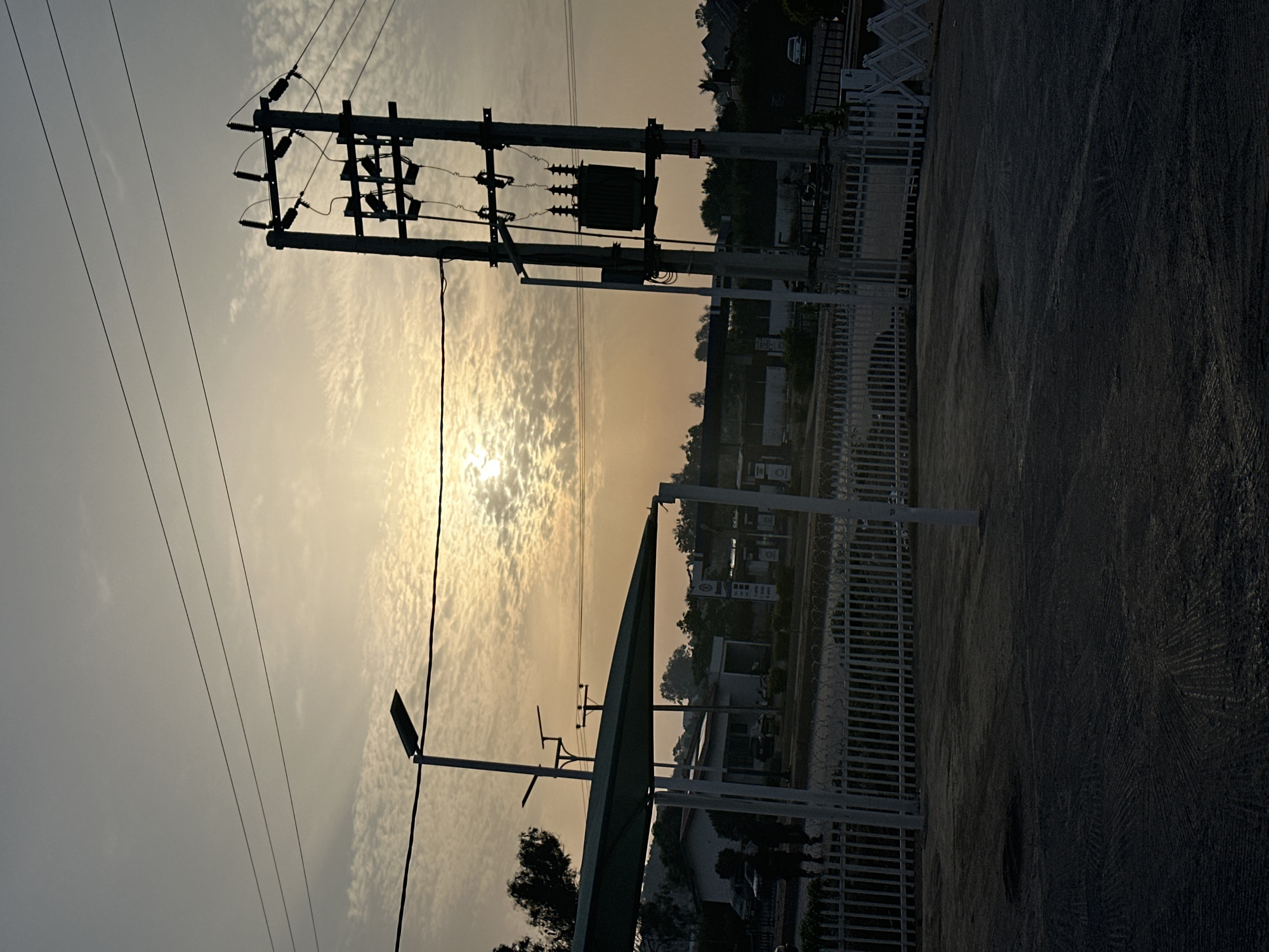 The image shows a view towards the horizon at dusk or dawn, with the sun partially obscured by clouds. In the foreground is an electrical power pole and transformer, with power lines extending across the sky. Below the power lines and pole is the roof of a covered structure, possibly a bus shelter or a similar facility. To the right of the structure is a white metal fence. The scene seems to be in an urban or suburban area, with buildings and trees visible in the distance. The location is noted as Bukuru, Nigeria. No people are directly visible. The overall impression is of a quiet moment in an urban environment with electrical infrastructure dominating the view of the skyline.