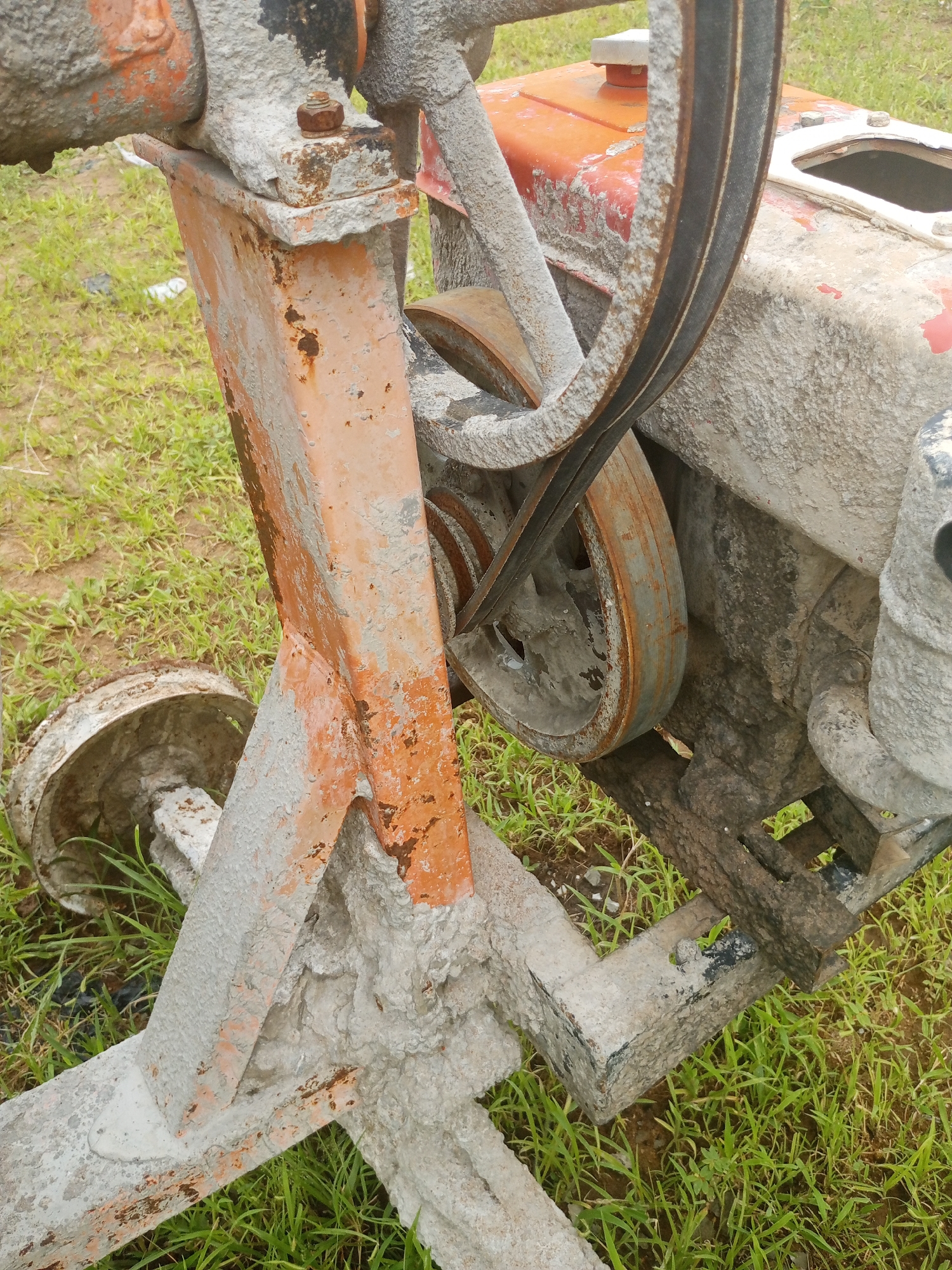 This image shows a close-up, outdoor view of a concrete mixer machine on a patch of green grass. The machine appears to be made of metal and has a significant amount of dried concrete caked onto its surfaces, indicating recent use. The orange-painted metal frame is rusted in places, and a black rubber belt is visible around two pulleys. The ground around the machine is covered in grass and dirt. The image does not show any people or specific landmarks to identify a precise location within Jalingo, Nigeria.