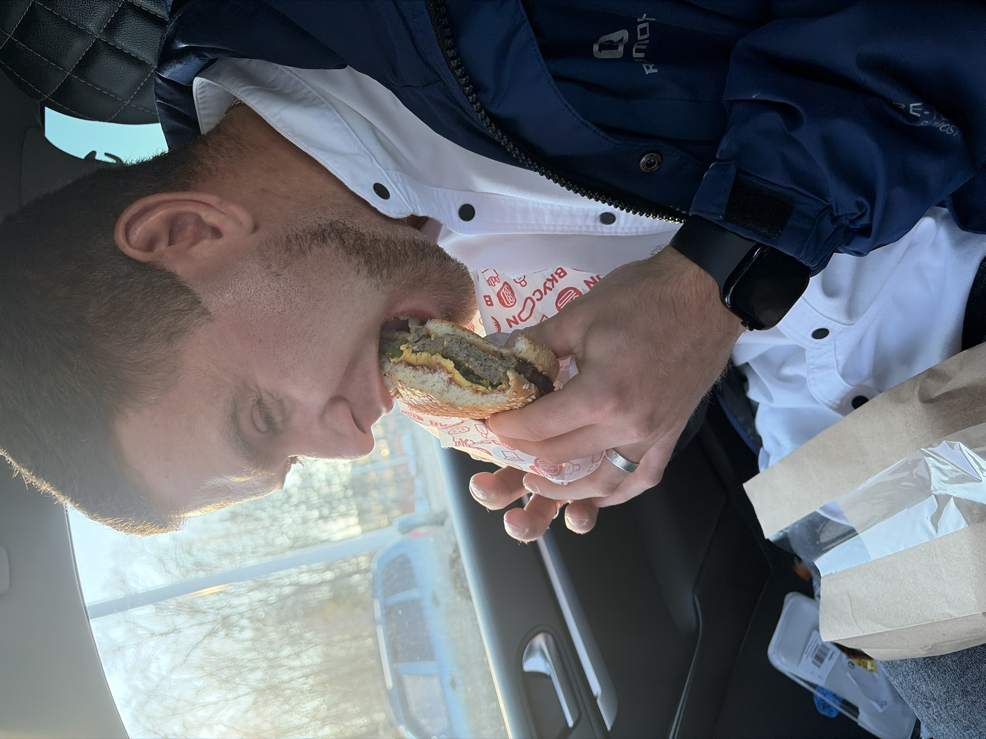 A man with short brown hair and a beard is seated inside a car, taking a bite out of a hamburger. He is wearing a white shirt, a navy jacket, and a black smartwatch on his left wrist. The burger is wrapped in a paper wrapper with red and white text. The car's interior is visible, including the dashboard, seat, and a brown paper bag. The background visible through the car window appears to be an outdoor environment with trees and possibly other buildings. There is no indication of location in the city of Votkinsk, Russia.