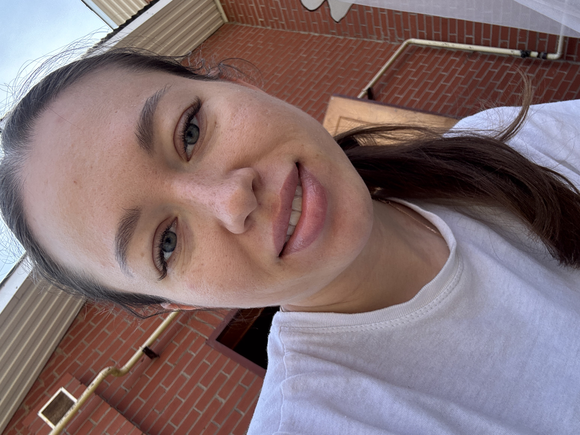A woman with fair skin, dark hair pulled back, and wearing a white t-shirt takes a selfie. The background includes a brick wall and eaves suggesting a building in Ферма, Russia.