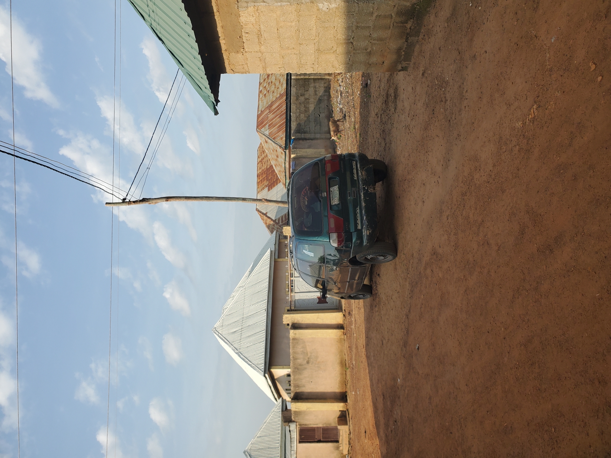 In Winters, United States of America, this outdoor scene depicts a dirt road in front of several buildings. A dark green minivan is parked on the left side of the road, angled towards the viewer. The minivan has a license plate with the characters "AXV 788 L". In the background, there are buildings with corrugated metal roofs, some appearing to be under construction or in disrepair. One building on the right is made of concrete blocks. A wooden utility pole with wires stretching across the sky stands behind the minivan. A light-colored animal, possibly a pig, is visible in the middle ground, walking away from the camera. The sky is partly cloudy with a clear blue sky visible between the clouds.