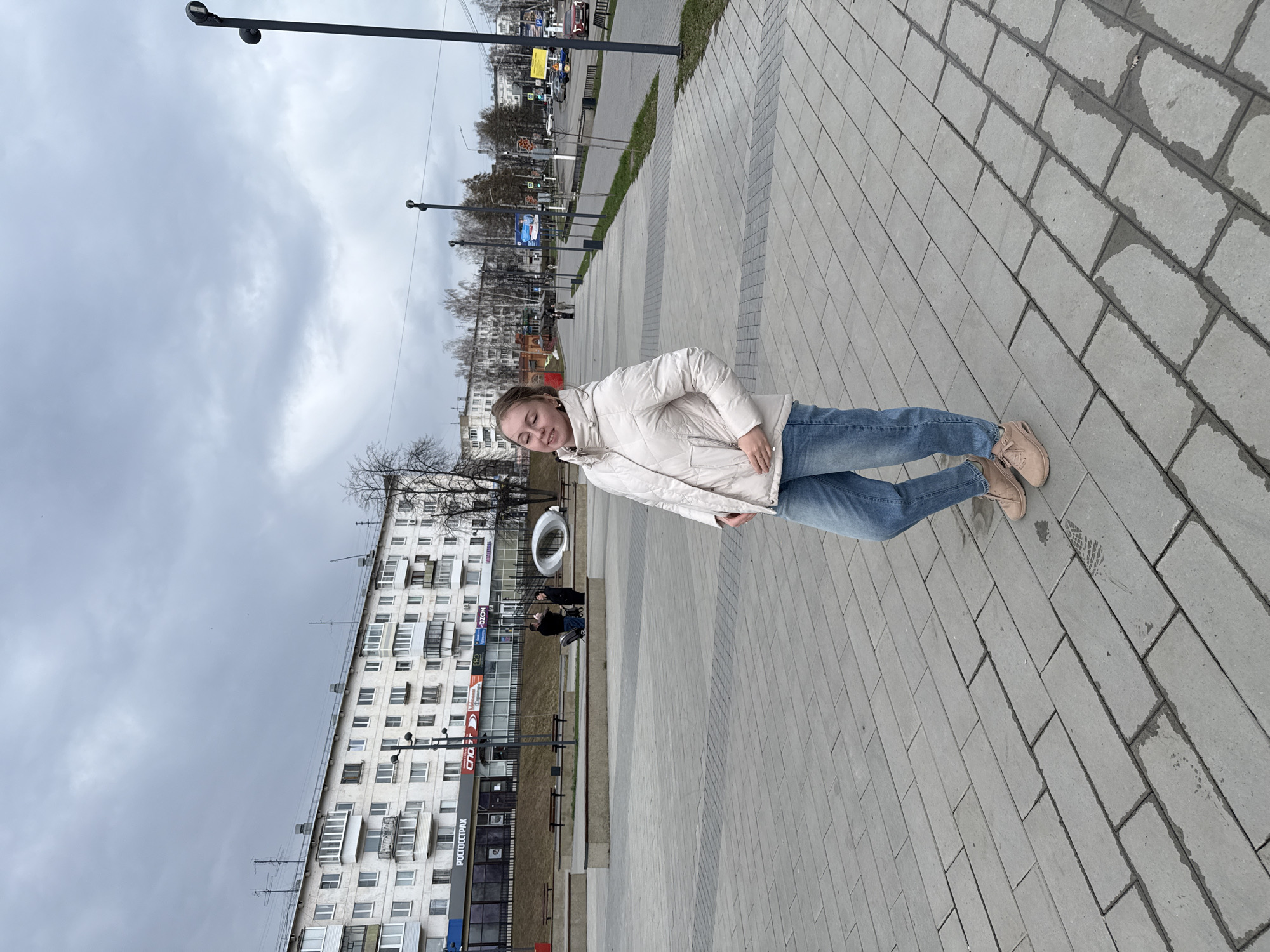 A woman stands on a paved sidewalk in an urban area. She wears a light-colored jacket and blue jeans. Behind her is a multi-story building with numerous windows. Streetlights line the road in the background, and a cloudy sky is visible.