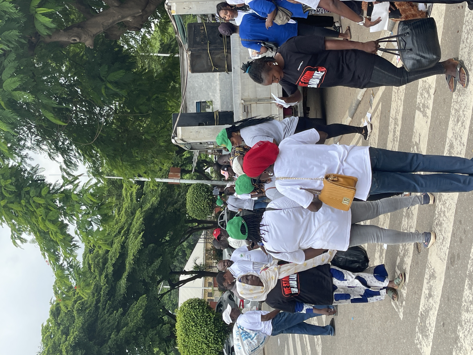 This media file captures a gathering of people in what appears to be an outdoor urban setting, possibly a street or plaza, in Abuja, Nigeria. The image is oriented vertically, with the subjects facing towards the right of the frame. The scene is filled with individuals, many of whom are wearing casual clothing. Some are dressed in white tops, while others are in black or blue attire. Several people are wearing caps, including green and red ones.
Among the objects visible are bags carried by some individuals, including a prominent black handbag and a smaller, quilted yellow crossbody bag. A large vehicle, possibly a truck or a stage, is present in the background, with speakers mounted on it, suggesting a potential event or rally. Trees with lush green foliage line the background, indicating an outdoor environment, and the lighting suggests it is daytime, possibly overcast.
The people are standing or walking, and their postures and expressions suggest a degree of focus. Some appear to be holding papers or looking downwards, possibly at documents or their phones. The overall atmosphere seems to be one of a planned gathering, with a collective presence rather than random individuals.
One notable detail is the presence of a white fabric bag slung over the shoulder of a person wearing a black t-shirt. This t-shirt has text on it, which is partially visible and reads "WE STAND FOR JABI LADI". Another visible text on a black t-shirt worn by a person on the left says "OMBDIKE". The ground shows a crosswalk pattern, further reinforcing the urban setting. The emotion conveyed is difficult to ascertain definitively from still images alone, but the organized nature of the group suggests a shared purpose.