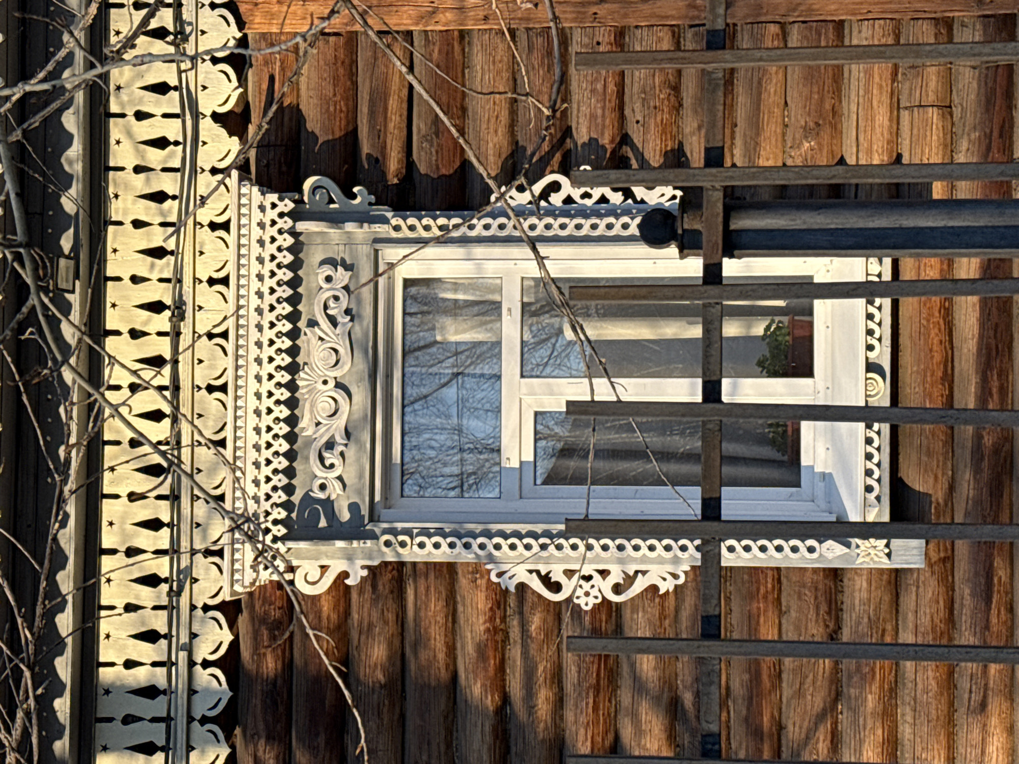 An eye-level, outdoor shot depicts a window of a wooden building, potentially located in Borzya, Russia, as indicated by the image request. The building's exterior is characterized by vertically arranged, rough-hewn wooden planks. The window is white-framed and adorned with intricate, decorative wood carvings along its top, bottom, and left side. Branches obscure portions of the window and building's facade. A dark, metallic structure resembling a ladder or scaffolding is positioned on the right side of the window, partially obstructing the view. The window reflects the sky, suggesting clear, daytime conditions.