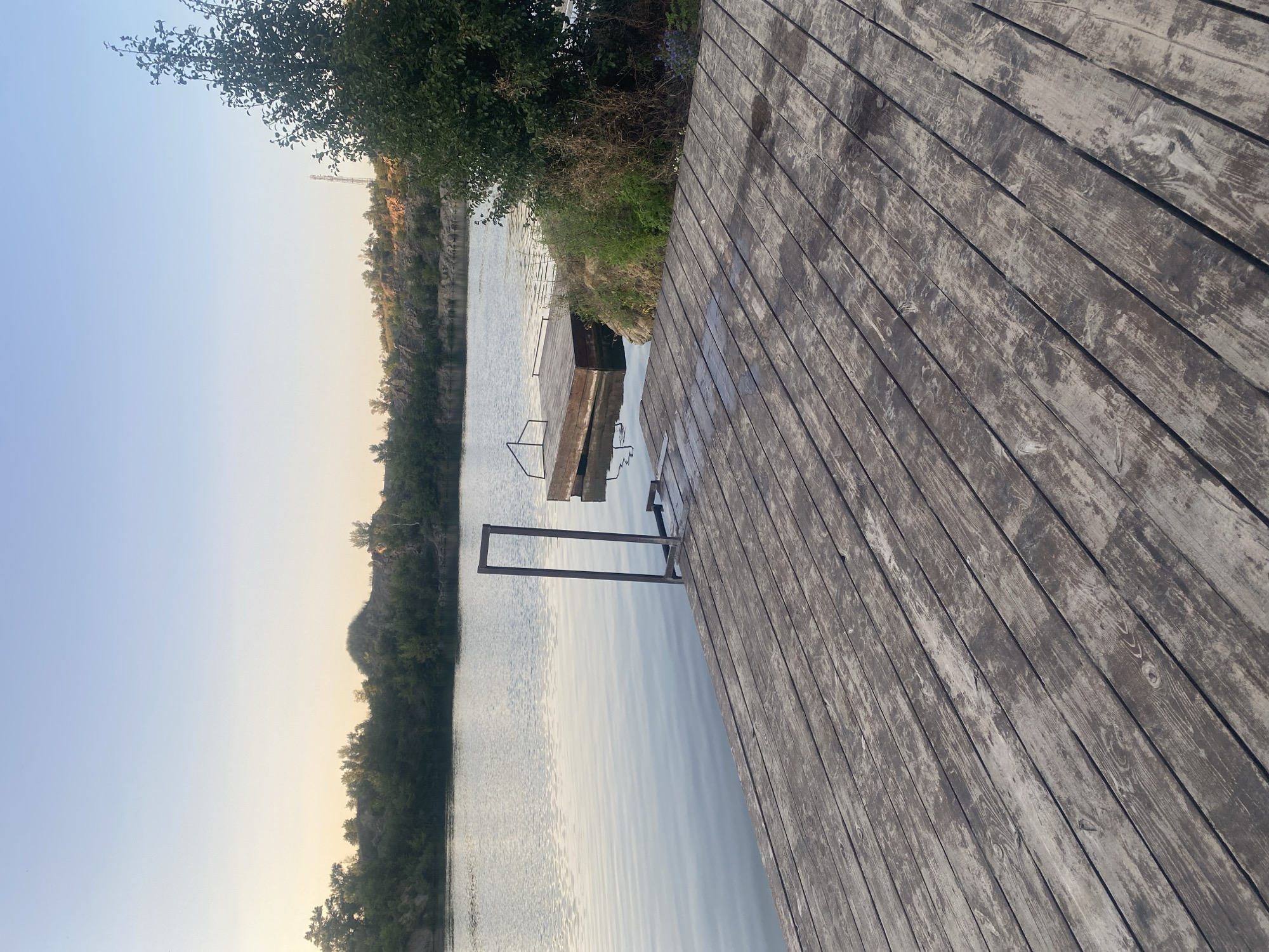 The image depicts a wooden pier extending into a calm body of water. The pier is made of weathered, grey planks laid parallel to each other. On the pier, there is a dark metal frame with a horizontal support at the bottom, seemingly a part of a structure for accessing the water. To the right, a smaller wooden platform floats on the water, also with a metal railing. The background shows a tree on the right side of the image, with its leaves obscuring part of the view. Beyond the water, there are gentle hills covered with sparse vegetation and trees. The sky is clear and pale blue, with a hint of yellow near the horizon, suggesting it might be early morning or late evening. The scene appears to be a tranquil natural setting, possibly a quarry or a lake area. The specific location in Dnipro, Ukraine, cannot be determined from the visual information alone.