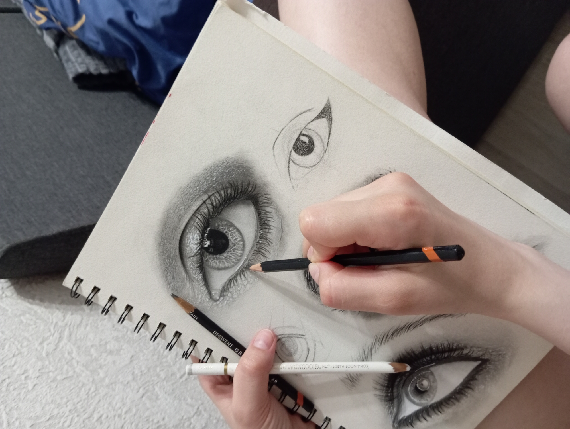 A person's hands are drawing multiple eyes on a sketchbook, in an indoor setting. The sketchbook is spiral-bound and open to a page with several graphite drawings of eyes. One eye is highly detailed, with lashes and shading. Another eye is sketched lightly, and a third is in progress. The person is holding two pencils: a white pencil in their left hand and a black pencil in their right hand, which they are using to draw. The background is out of focus, showing a textured wall and a dark piece of furniture. The lighting appears to be artificial.