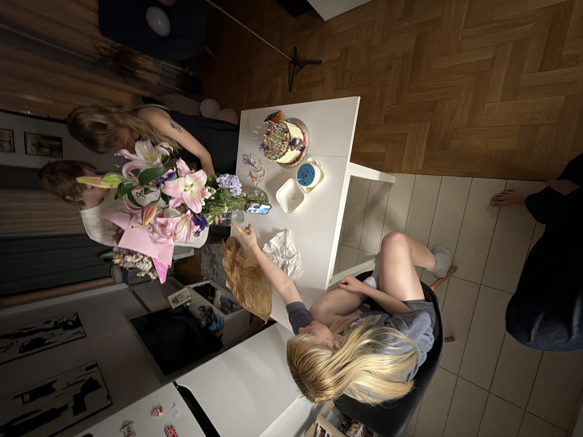 In a dimly lit room, likely a residential apartment in Vračar Urban Municipality, Serbia, three individuals are gathered around a white table, seemingly for a birthday celebration. On the table, a tiered cake with colorful frosting and lit candles sits next to a smaller cake with the number '21' displayed on it. A bouquet of pink lilies and purple flowers is arranged in a vase, and a mobile phone is propped up nearby. One person, seated, with blonde hair and wearing a grey t-shirt and shorts, points towards the cake. Two other individuals, one with blonde hair and wearing a black dress, and another whose face is obscured, are standing and looking at the bouquet. The room features wooden flooring, a refrigerator, and artwork on the wall. The atmosphere appears to be one of intimate celebration.