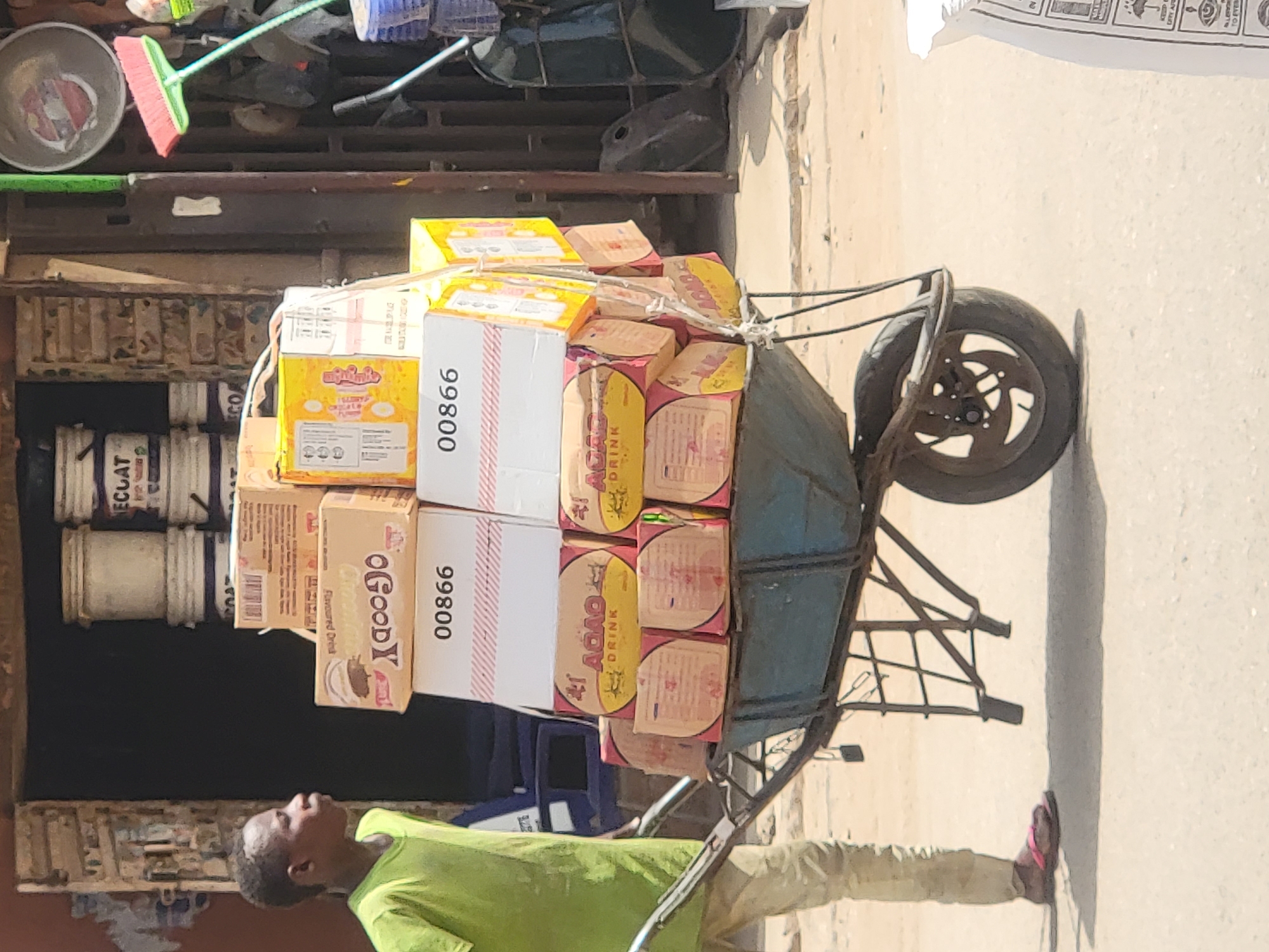 A man in a green shirt and khaki pants is pulling a blue wheelbarrow, which is loaded with multiple boxes of goods. Some of the boxes are labeled "OGOODY Flavoured Drink" and "#1 ADAO DRINK." Other boxes display the number "99800." The wheelbarrow is on a paved surface, likely a street or sidewalk. In the background, there's a building with various items stored, including paint cans labeled "ECLAT." The location appears to be a commercial or market area, suggesting the man is transporting goods for sale or distribution. The city of Maiburiji, Nigeria is mentioned in the prompt.