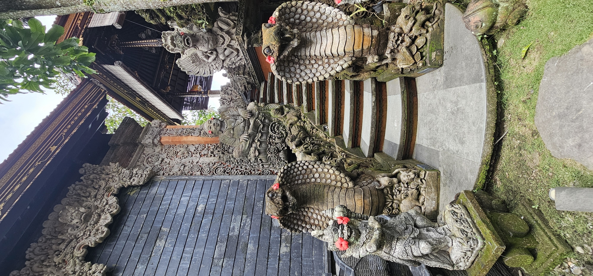 The image captures a temple entrance in the Ubud District of Indonesia. The architectural structure features intricately carved stone statues flanking a set of stairs. These statues depict cobras with red flowers placed near their heads, and other figures are present amongst elaborate stone carvings that adorn the temple facade. The stairs have light stone risers and darker treads. Moss and greenery grow on the stone elements, indicating age and exposure to the humid environment. A portion of a traditional Balinese roof is visible above the entrance. The ground near the stairs is covered with grass. The lighting is soft and natural, suggesting overcast conditions.