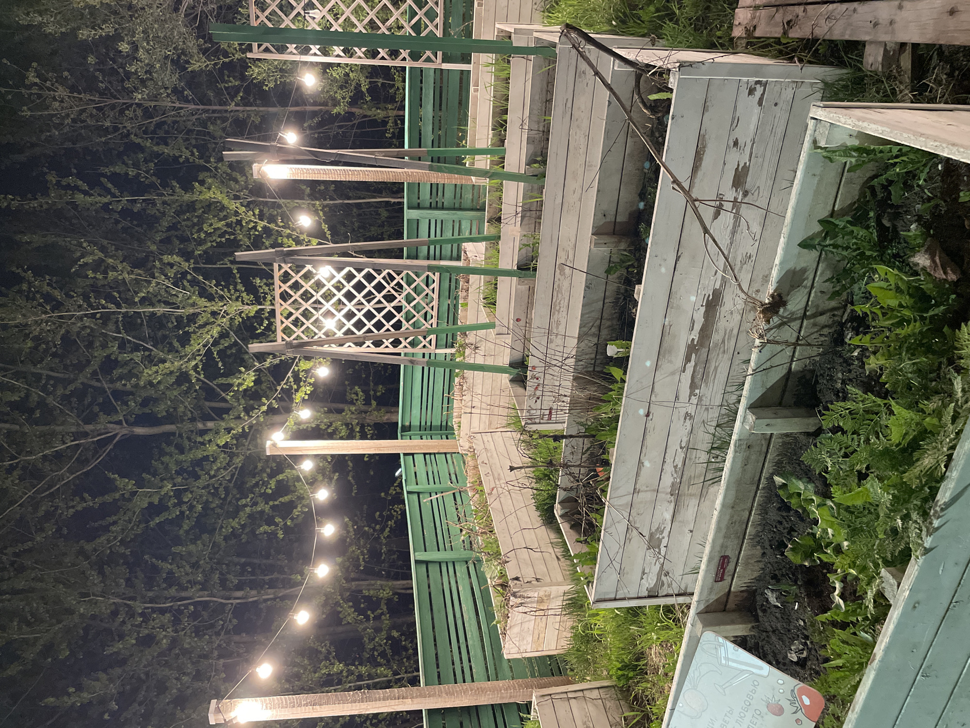 The image shows an outdoor space, likely a garden or patio, at night. String lights illuminate the area. There are wooden structures, including raised garden beds and what appears to be a green-painted fence or screen with lattice sections. Plants are visible in the raised beds. The location is specified as Kazan, Russia. No people are visible.
