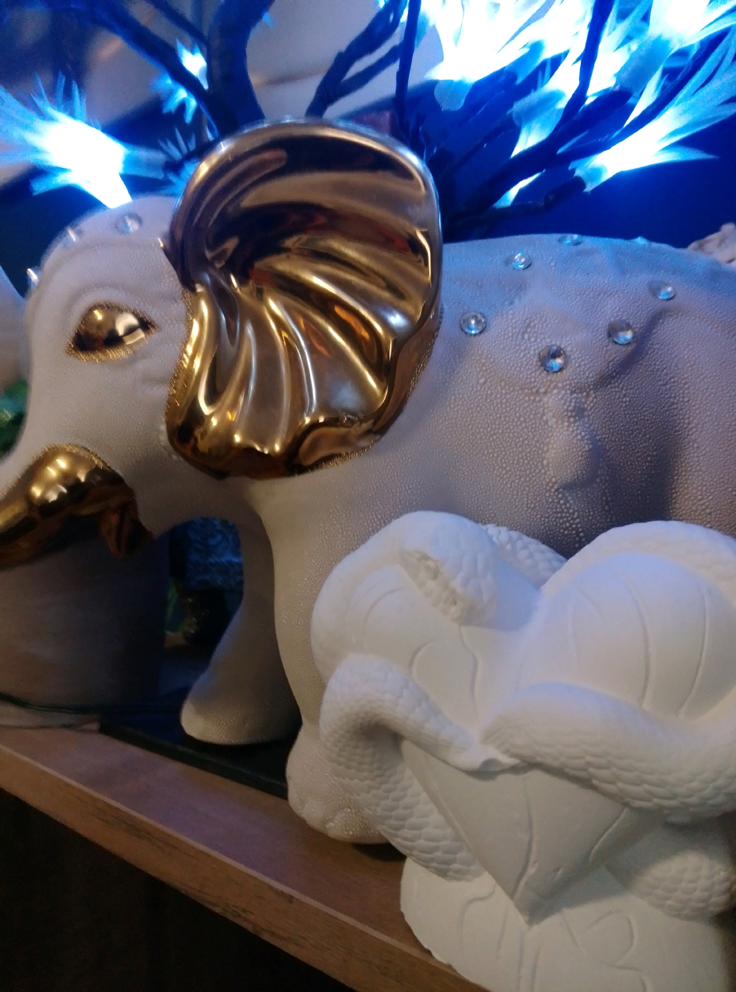 This is a close-up, low-angle, vertical photograph of two decorative figurines on a wooden shelf. The figurine in the foreground is a white, matte sculpture of a heart-shaped object entwined with a snake. The snake's scales are detailed, and the heart shape appears to be formed from petals. The figurine in the background is a white elephant statue with gold accents on its ears and tusks. The elephant's skin has a textured, pebbled appearance, and several small, clear rhinestones are embedded in its body. The elephant's eyes are also outlined in gold. Behind the elephant, a dark, branch-like structure is illuminated by bright blue lights, creating a dramatic and somewhat abstract background. The lighting is dim overall, with the blue lights and the gold accents being the brightest elements in the scene. The image appears to be a still life of decorative items, possibly in a home or shop setting. There are no people or animals present. The location in Wedel, Germany is not ascertainable from the image.