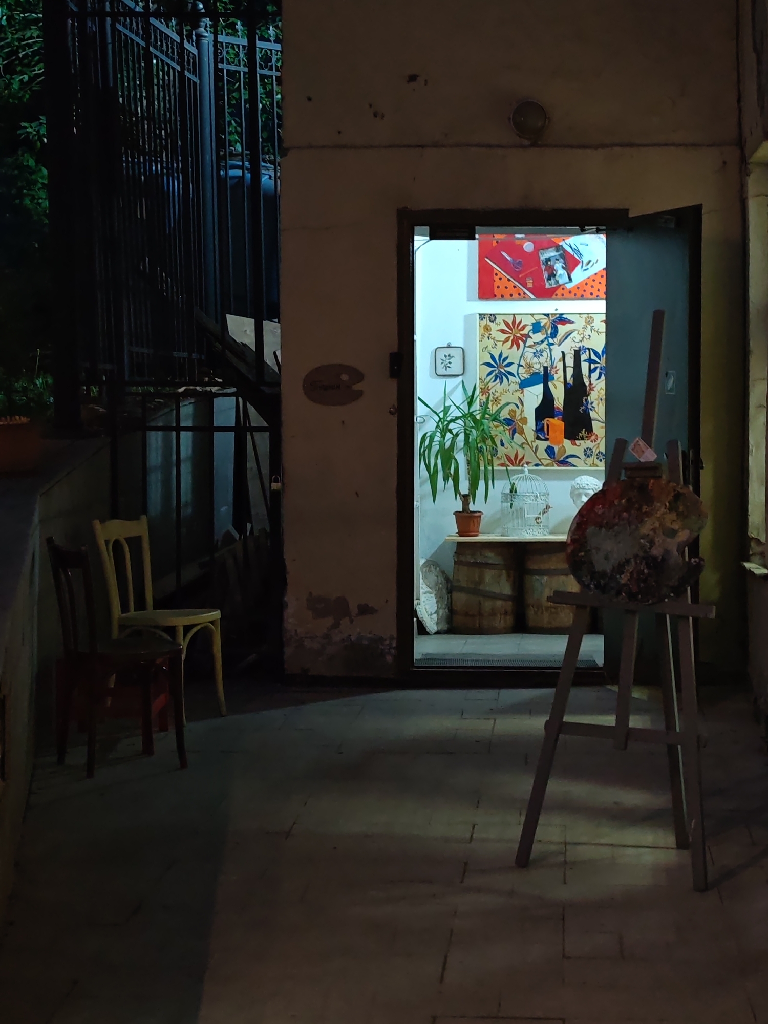 The image shows an open doorway to a brightly lit room, likely a studio or gallery, adjacent to a dark exterior space. The location is suspected to be in Moscow, Russia.
The doorway reveals the interior of the room. Inside, two framed pieces of art hang on the wall: one is orange with red and white details at the top of the frame, and another larger floral piece below. A potted plant is positioned on a rustic wooden table, constructed from two barrels, along with a white birdcage and a partial bust sculpture.
In the foreground, to the right of the doorway and outside, is an easel holding a paint palette loaded with various colors.
To the left of the door and outside, there are two wooden chairs: one with a yellow back, and the other a reddish-brown hue. To the left of the chairs is a metal gate or fence, suggesting an enclosed area or yard.
The overall scene appears to be a quiet, possibly creative space, juxtaposing the dimly lit exterior with the brightly lit interior. The lighting suggests it might be evening or night. The environment gives the impression of a rustic or vintage setting, possibly an artistic enclave in an urban area.