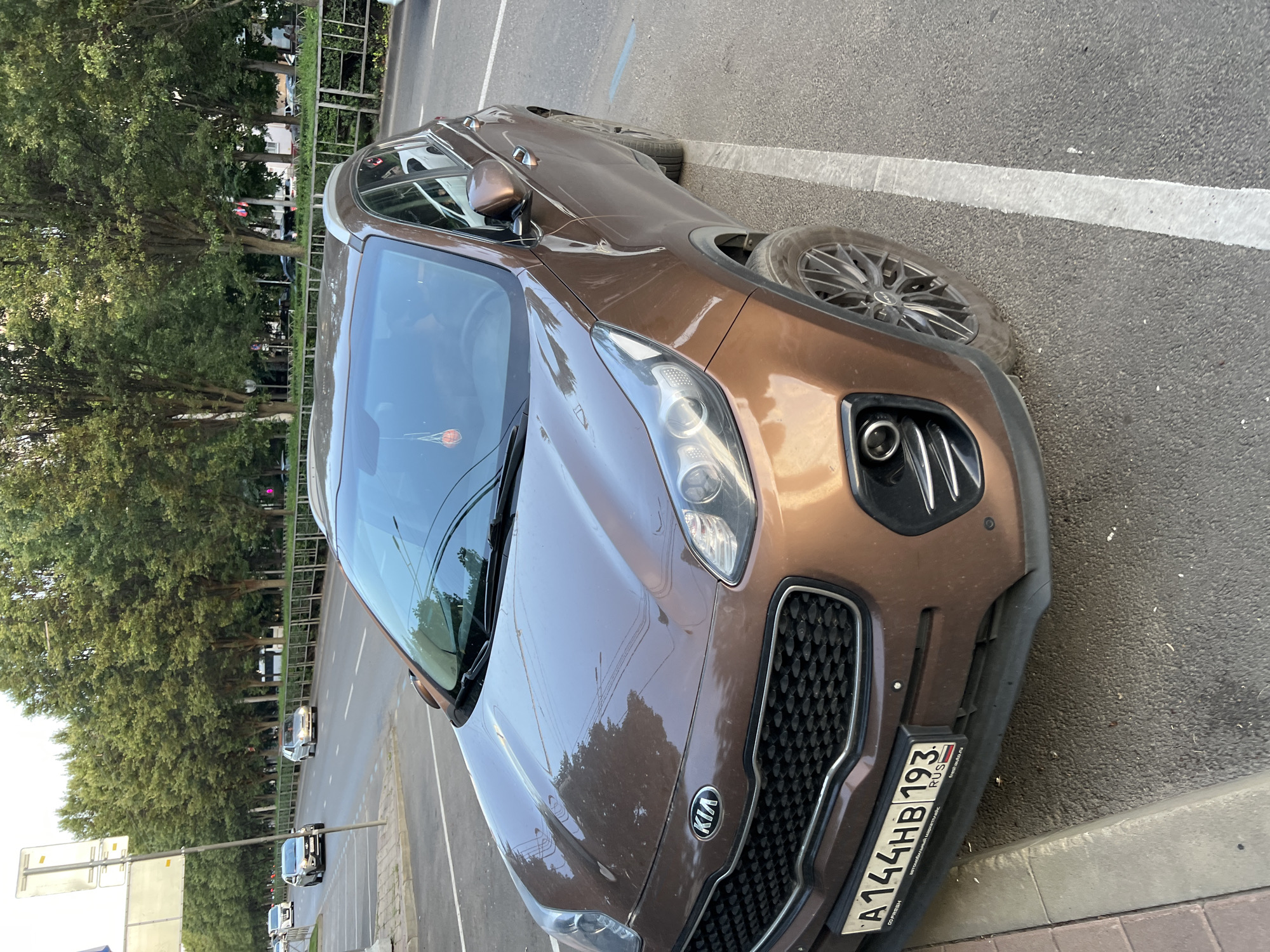 A brown 2017 Kia Sportage SUV is parked at an angle on a city street in Krasnodar, Russia. The vehicle is in the foreground, facing the right side of the frame. The license plate reads "A144HB 193 RUS". The street is paved and marked with white parking lines. To the left of the car is a sidewalk with a curb. In the background, there are trees lining the street, and further back, buildings and other cars can be seen. The sky is overcast.