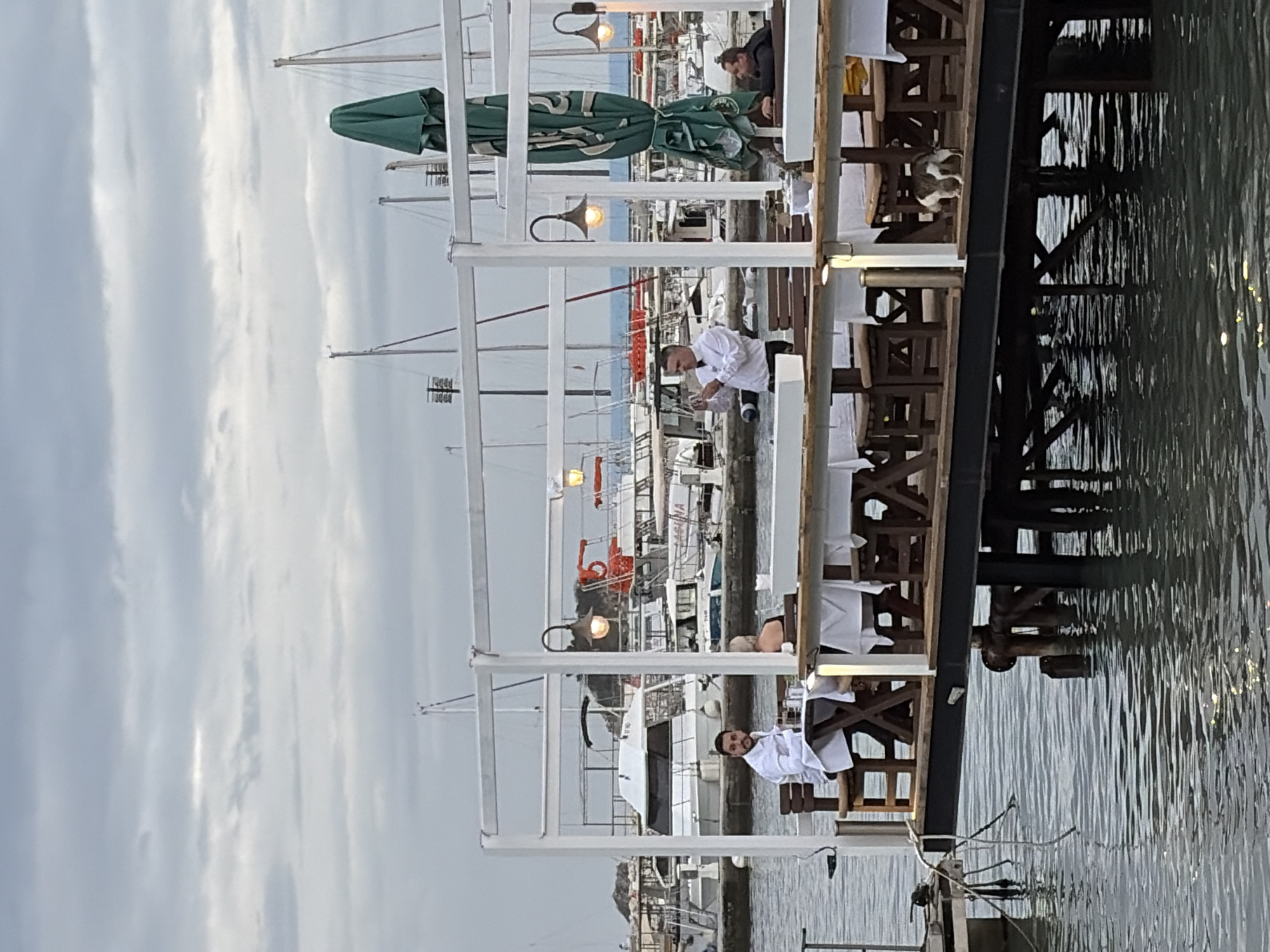 The image captures an outdoor restaurant scene in Budva, Montenegro. The restaurant is positioned on a pier or dock extending over the water, with a view of the sea and various docked boats. White table linens cover the tables, and the restaurant structure features white architectural elements, including railings and beams.
Several individuals are visible: two appear to be waiters dressed in white, engaged in setting tables; one is standing and one is sitting. Another person sits under a green umbrella near the back of the restaurant.
The foreground shows the water, which appears calm with visible reflections. The sky is overcast, suggesting an overcast day.