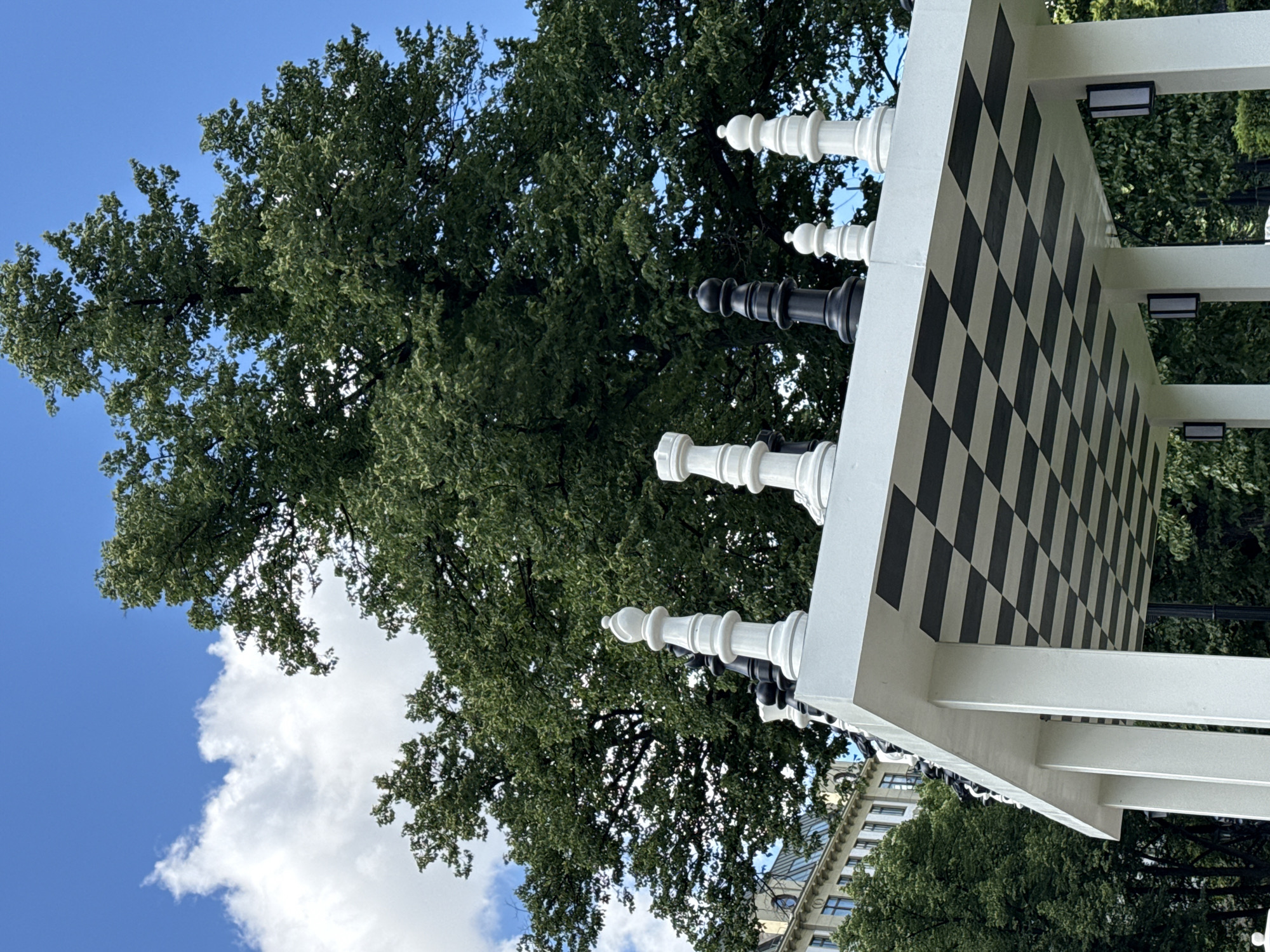 In Moscow, Russia, a large outdoor chess set is featured prominently in the foreground. The set, with its oversized pieces, rests on a white structure with a checkered black and white underside. The chess pieces are positioned on the top surface of the structure. A large, leafy green tree dominates the background, with a clear blue sky and scattered white clouds above. To the left, a building with multiple windows is partially visible. The scene appears to be a public space, likely a park or square, given the presence of the chess set and the surrounding greenery. There are no people visible in the image.
