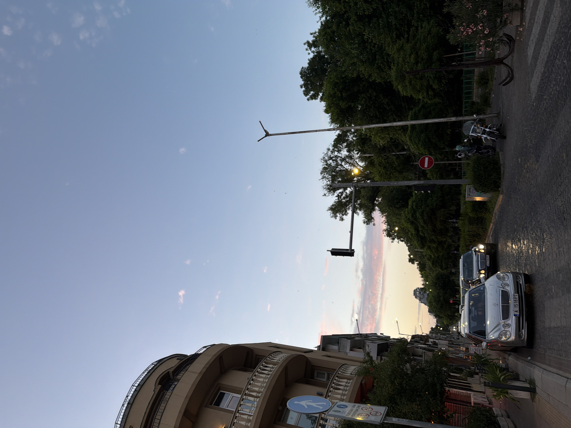 The image depicts a street scene in Burgas, Bulgaria, during what appears to be dusk. The sky is a soft blue with streaks of pink and orange clouds, indicating the sun is setting.
The foreground features a street with parked and moving vehicles. A silver Mercedes-Benz sedan with the license plate "A 1518 PT" is prominently visible on the right side of the road, facing away from the camera. Behind it, another vehicle, possibly a van or SUV, is also visible. Two motorcycles are parked on the left side of the street, near a decorative anchor-shaped sculpture.
On the left side of the image, a building with a curved facade and ornate balconies is partially visible. The building has a light-colored exterior and appears to be a residential or commercial structure. Street signs are also present, including a pedestrian crossing sign and what seems to be a "no entry" sign with a red circle and horizontal bar.
The background is dominated by lush green trees, which line the street and form a dense canopy. A traffic light is visible in the distance, suggesting an intersection. The overall atmosphere is calm and serene, characteristic of an early evening in a city. The lighting suggests it's either early morning or late evening.