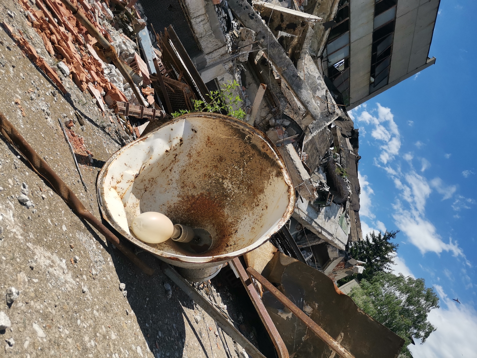 The image depicts a dilapidated building in Zhivkovo, Bulgaria, likely the result of destruction or decay. In the foreground is a rusty, white, megaphone-shaped object containing a light bulb. Debris, including bricks and metal fragments, litters the ground around the megaphone. The background shows the partially collapsed building, with exposed concrete and twisted metal. The sky is visible above the building, featuring blue patches and scattered clouds.