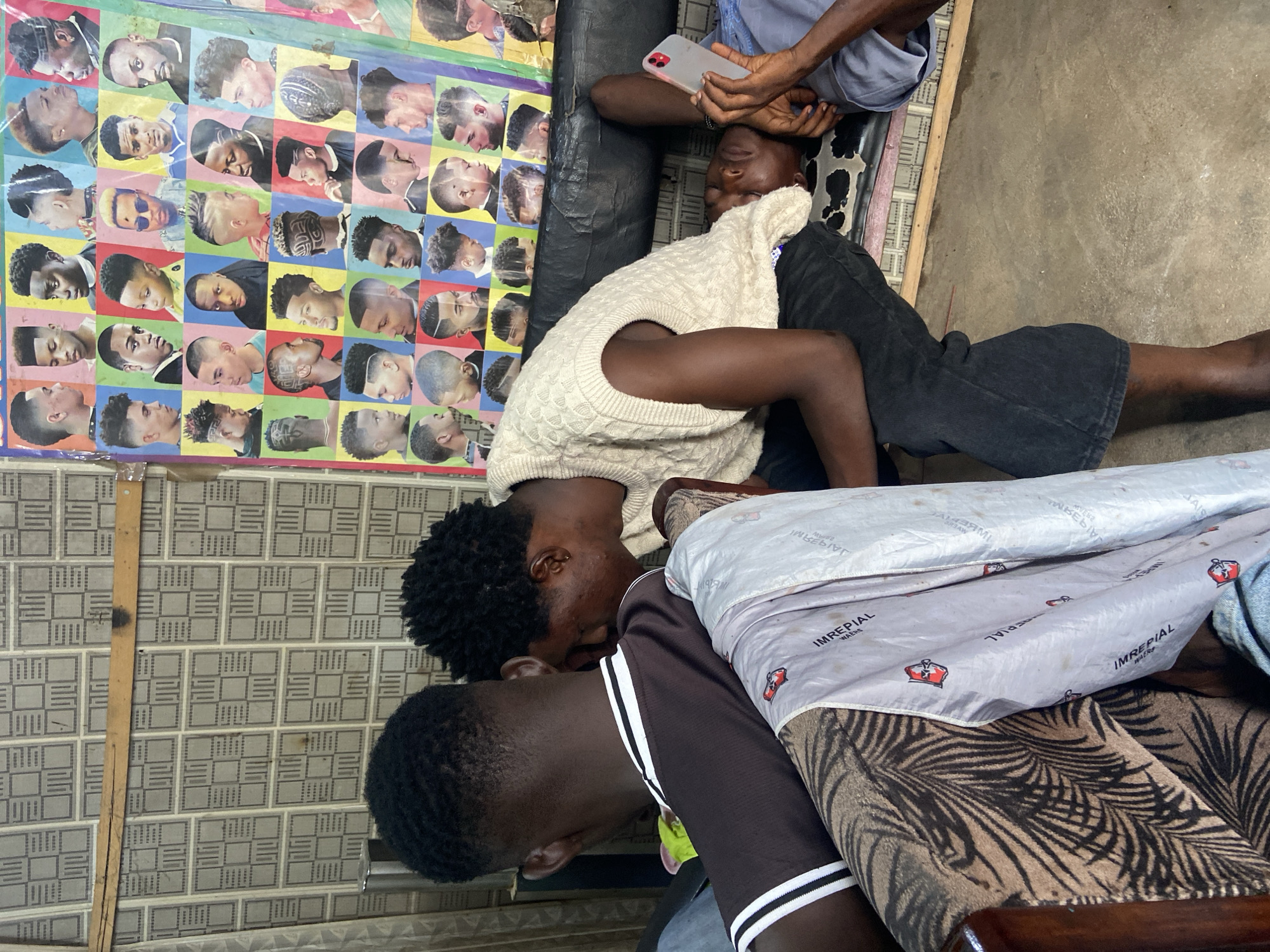 The image shows three young Black men in what appears to be a barbershop in Kuson, Nigeria. Two of the men are lying down, possibly resting or having their hair cut. The man in the foreground, wearing a dark brown shirt with white stripes on the sleeves, is lying on his back with his head resting on a patterned blanket. His eyes are closed, and he seems relaxed.
Another man, wearing a light-colored, cable-knit sleeveless sweater over a dark shirt, is lying next to him, also with his head down. His eyes are closed as well. He is holding a pink smartphone in his right hand.
A third person, whose face is partially visible, is seated or leaning over the other two, wearing a blue shirt. Their hands are holding the phone that the man in the sweater is touching.
Behind the men, a colorful poster displays numerous portraits of men with various hairstyles, typical of a barbershop. The walls appear to be tiled with a gray, patterned design. The floor is concrete.
The scene suggests a relaxed atmosphere, possibly during a break or after a service. The lighting indicates daytime, with no specific weather conditions visible. The presence of the barbershop poster and the haircuts on display clearly establish the context of a place where hairstyling services are offered. The overall impression is one of community and everyday life in a Nigerian barbershop. No visible text is prominent enough to be transcribed, other than what might be on the poster or the blanket, which is illegible.