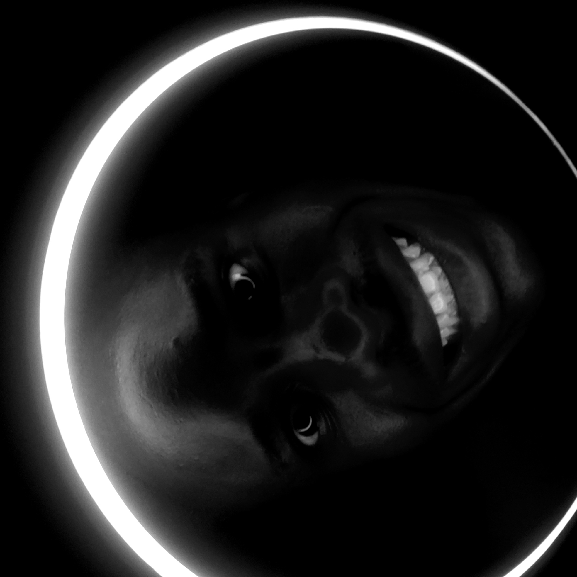 A grayscale image depicts a person's face framed by a luminous ring. The person is smiling, and their face is dark against a black background. The ring is a bright white arc encircling the face. There is no contextual information to ascertain its location in Narayi, Nigeria. The scene is minimalist, focusing on the contrast between the dark face and the light ring.