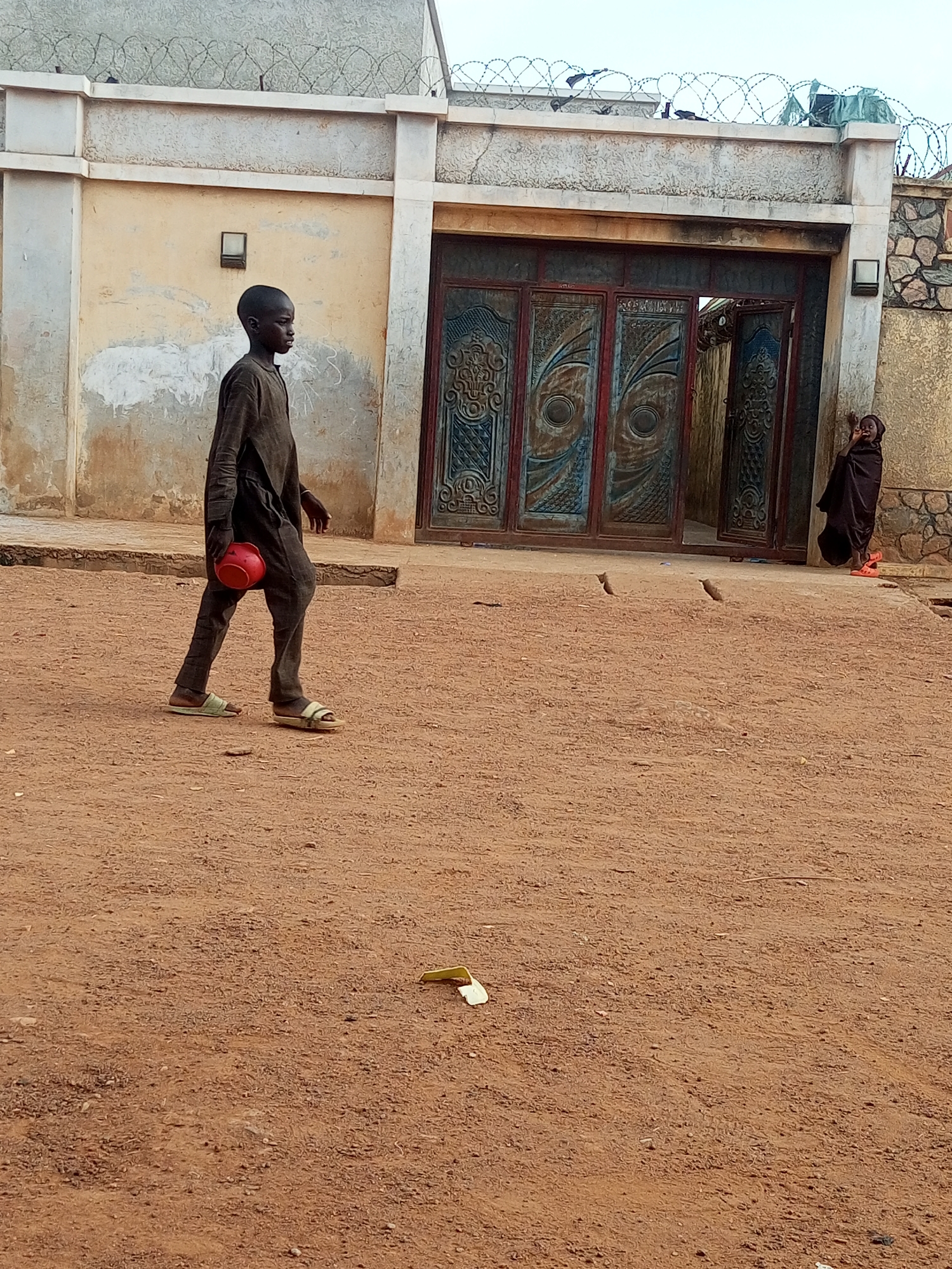 A young boy walks left across a dusty, reddish-brown yard. He wears a dark, loose-fitting outfit, sandals, and carries a red container in his left hand. Behind him is a building with a faded yellowish-beige facade and a large metal gate with intricate blue and red designs. Another child, draped in a dark garment, stands near the partly open gate, holding a hand to their face. Above the building's wall is barbed wire. The yard contains scattered debris, including a yellow object on the ground. Two small birds are visible near the building's entrance. The location is Maiburiji, Nigeria.